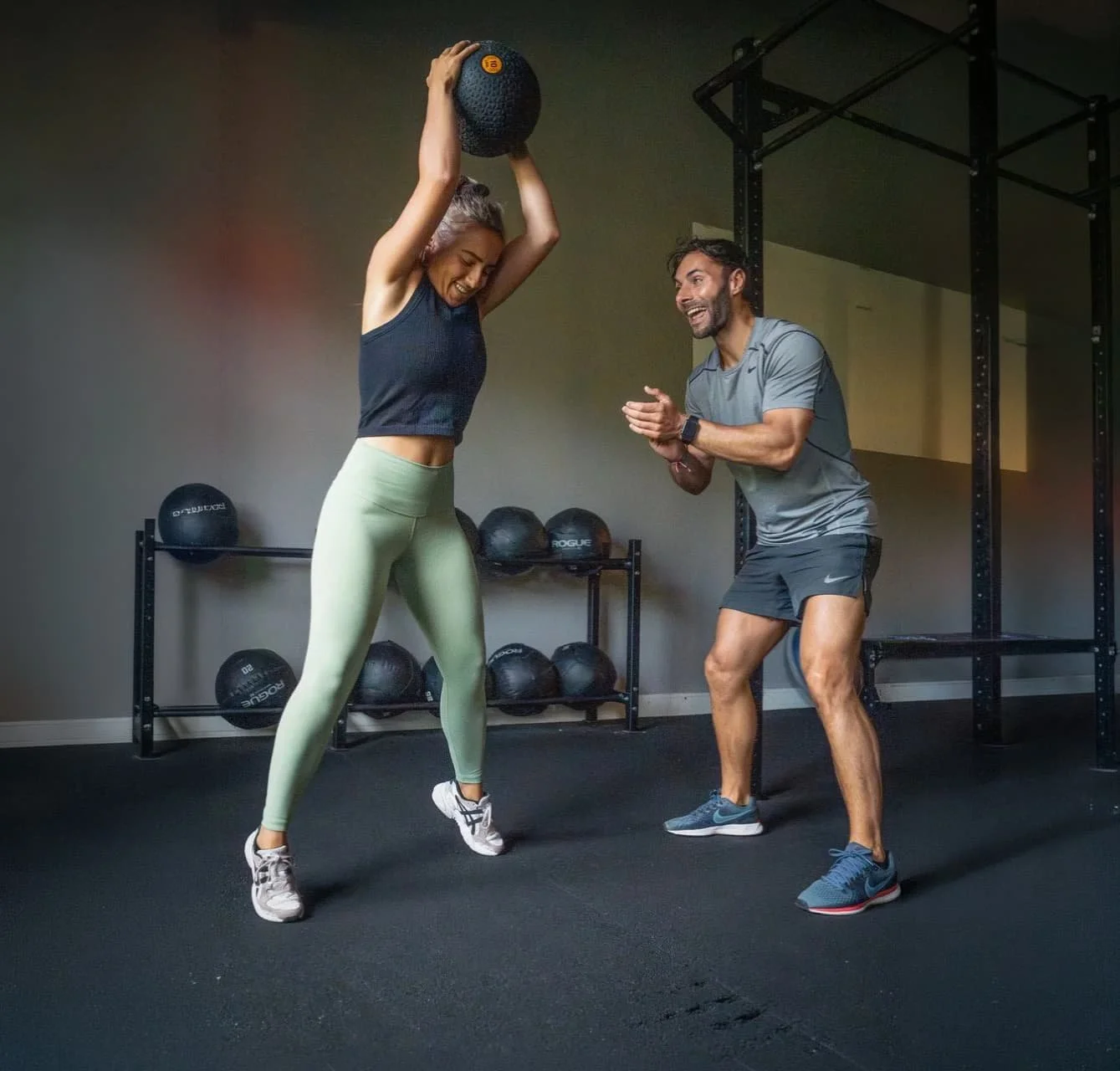 Twee mensen in de sportschool, een vrouw die een medicijnbal overhead houdt en een man die haar aanmoedigt.