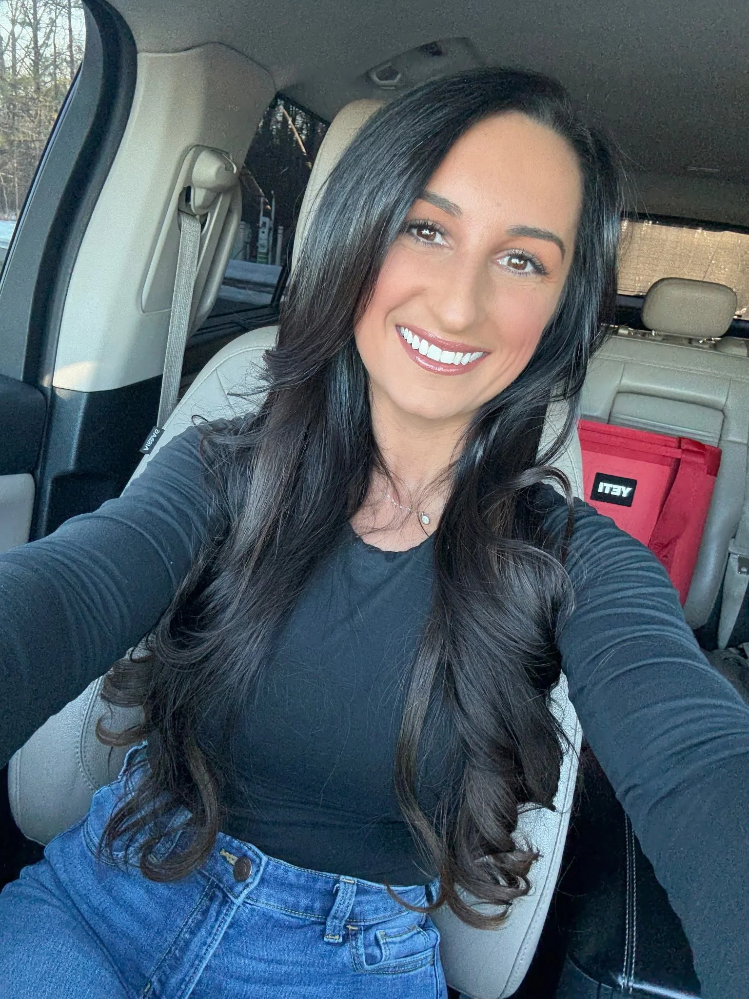 A woman with long dark hair smiling, seated in the back of a car, wearing a black top and blue jeans, with a red bag in the background.