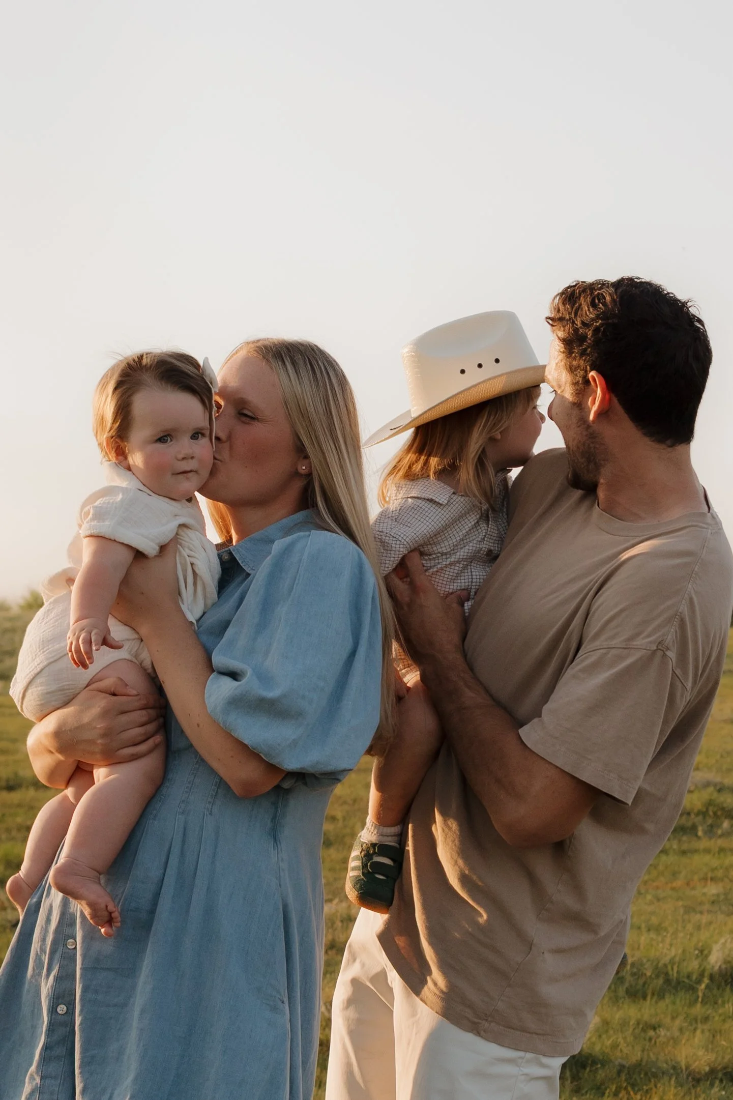 A family of four outdoors during sunset, with two adults each holding a child, sharing a kiss with their children, in a natural grassy field.