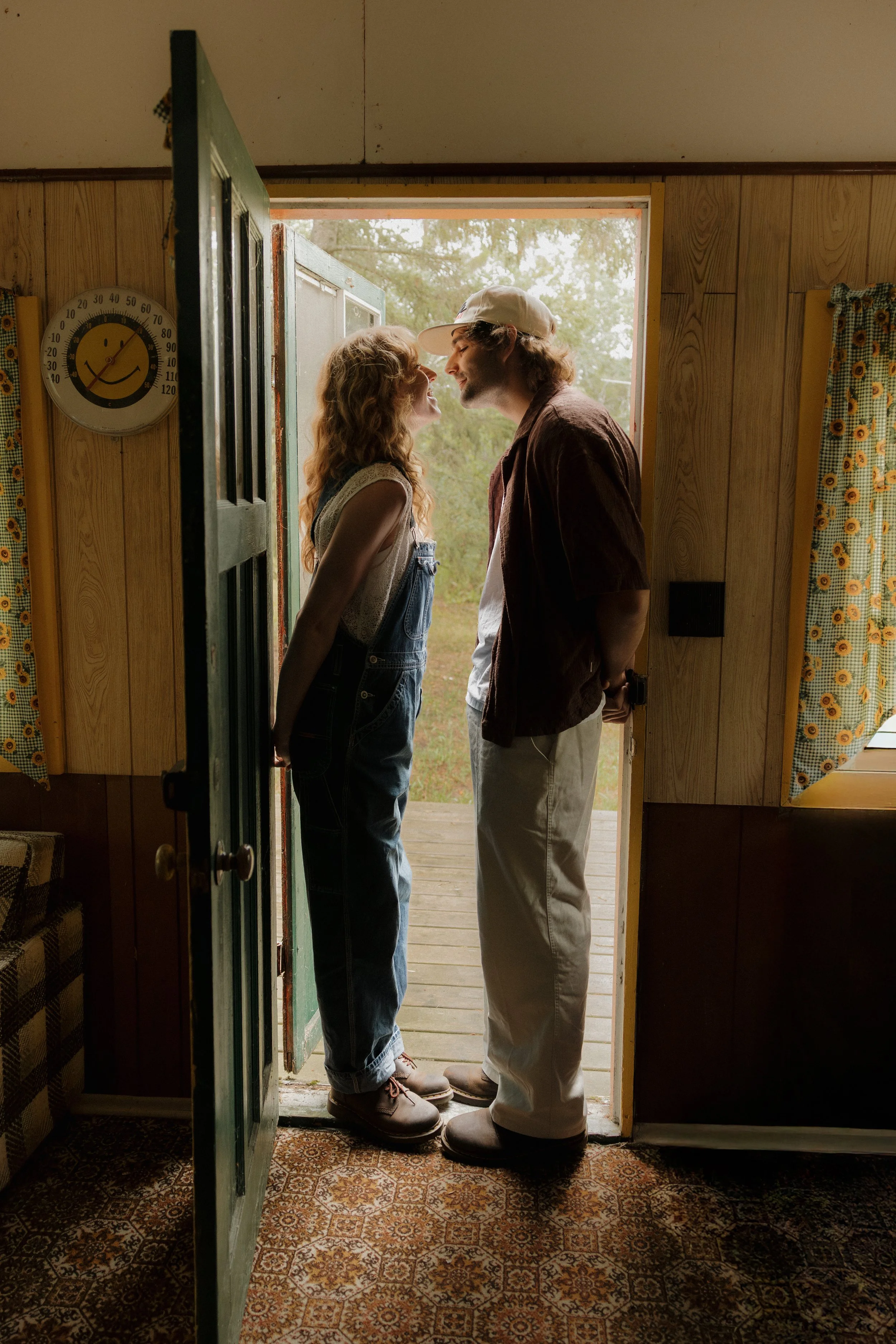 A young couple standing at a doorway, leaning in close with noses nearly touching, smiling in a rustic, cozy room with wood-paneled walls and patterned curtains.