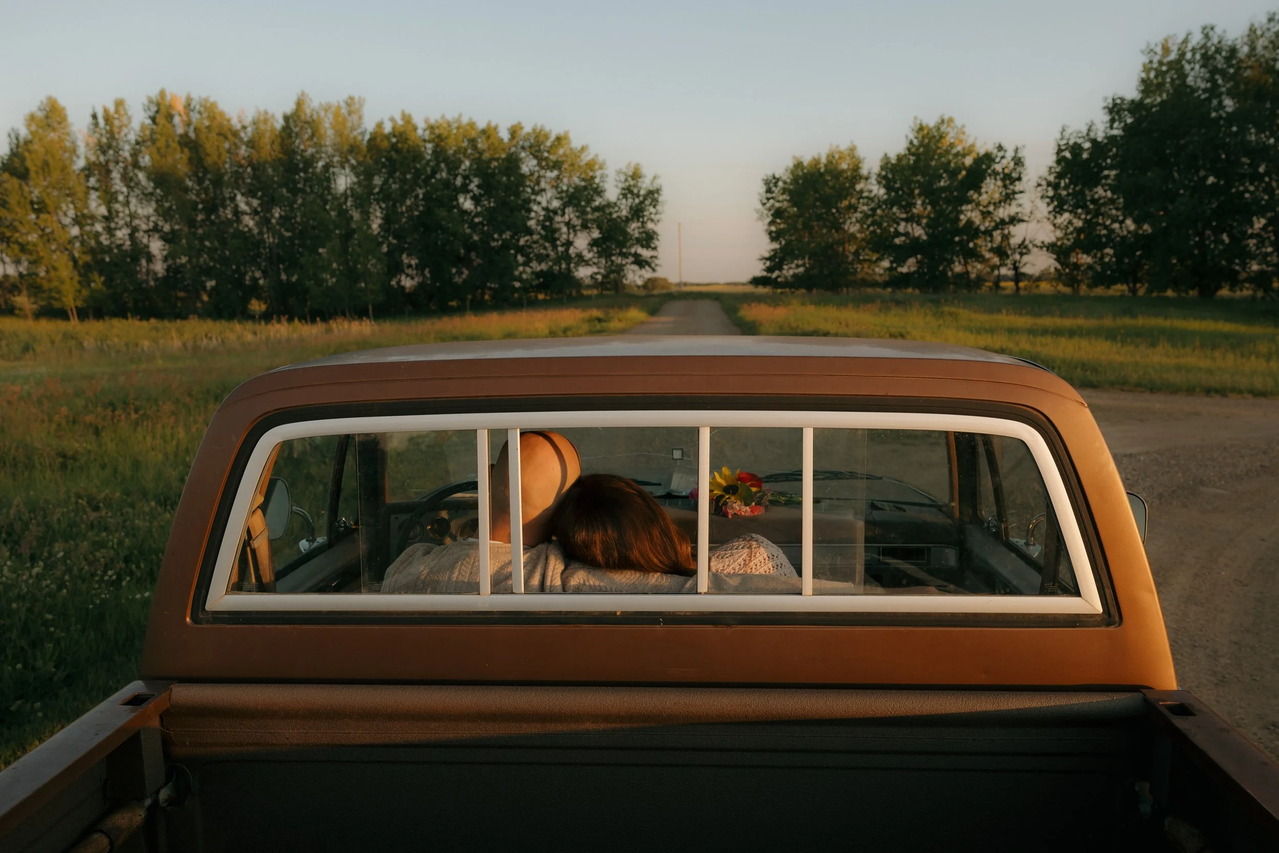 A truck on a rural dirt road with a couple resting under the sunset. The couple is sitting in the truck's cab, leaning their heads against each other, with a bouquet of flowers on the dashboard.