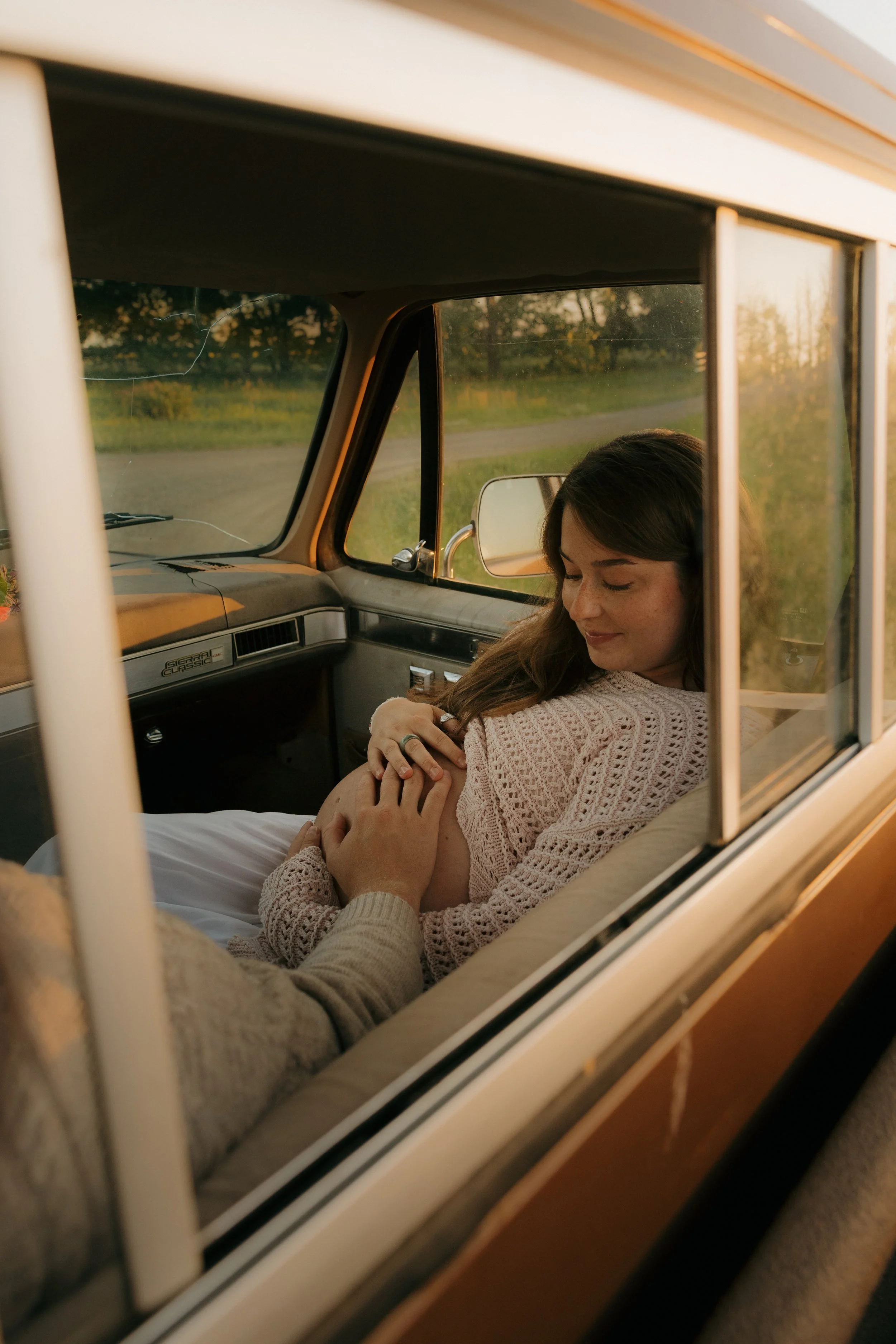 A woman sitting inside a vintage van, smiling and gently holding her pregnant belly while looking down, with another person’s hand resting on her stomach. The scene is set during sunset with greenery in the background.