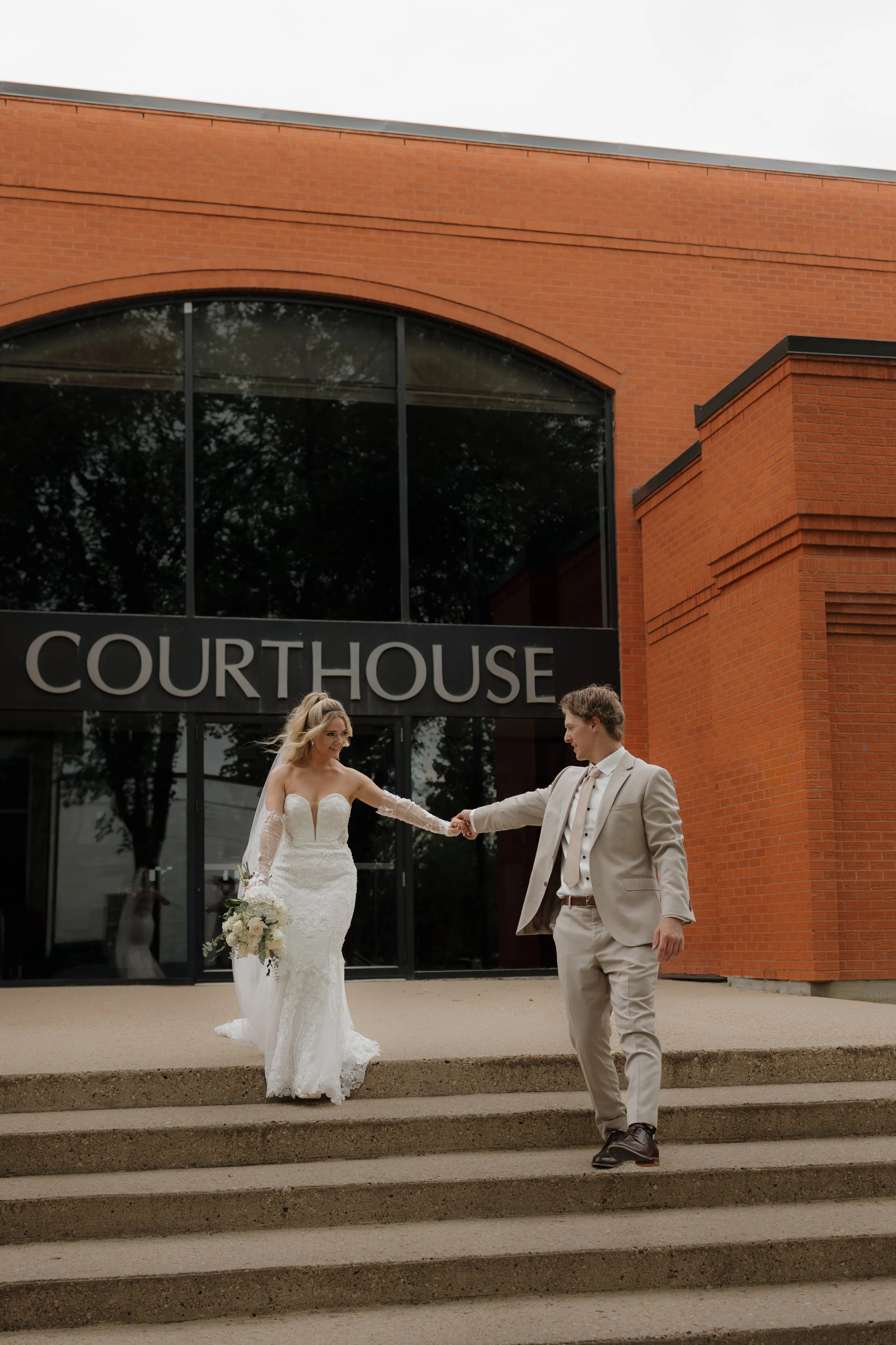 A bride and groom holding hands on the steps outside a courthouse building, with the bride holding a bouquet of flowers.