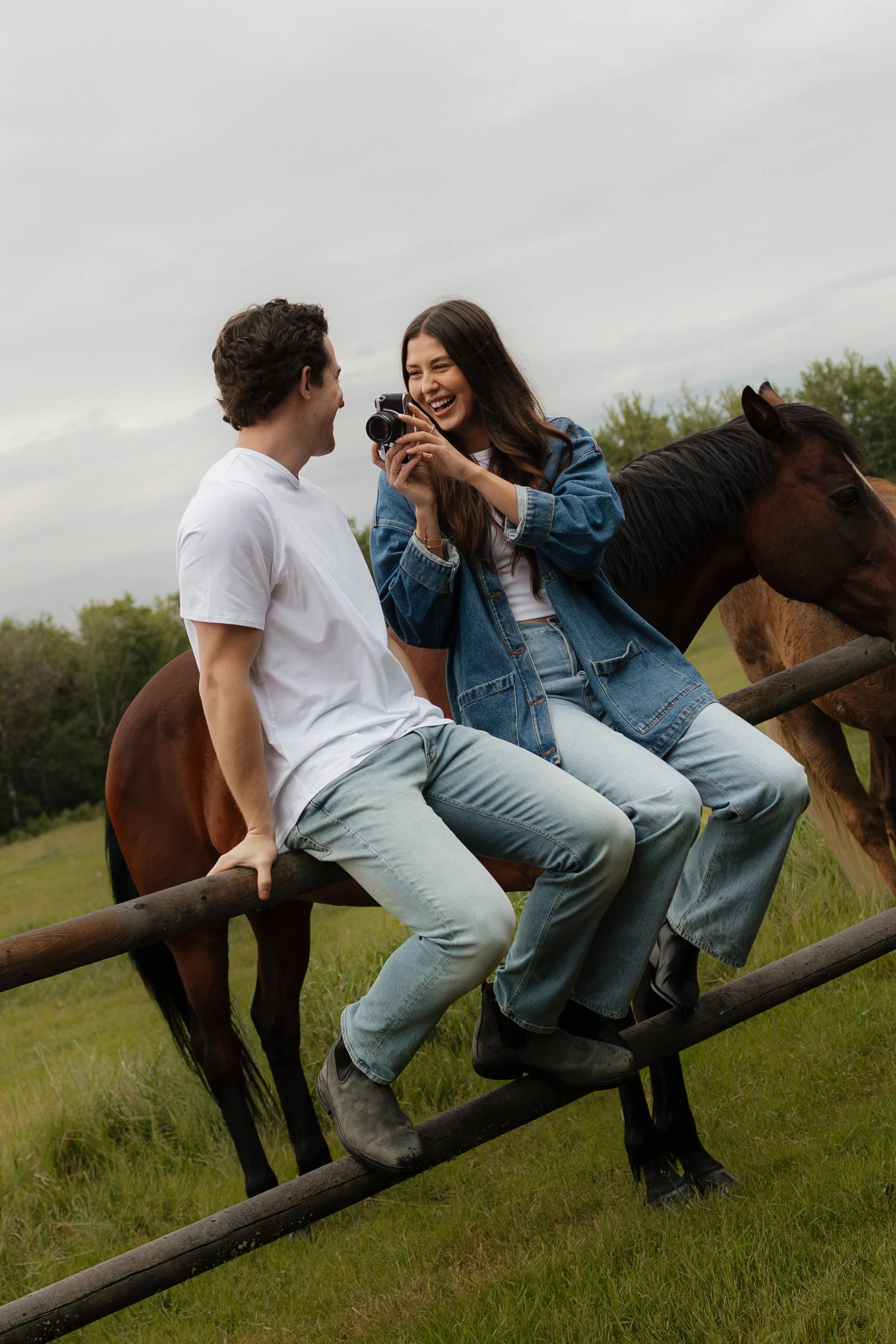 A young couple is sitting on a wooden fence in a field, sharing a joyful moment. The woman is holding a camera, and both are smiling and laughing. There are horses nearby, and the sky is cloudy with a backdrop of green trees.