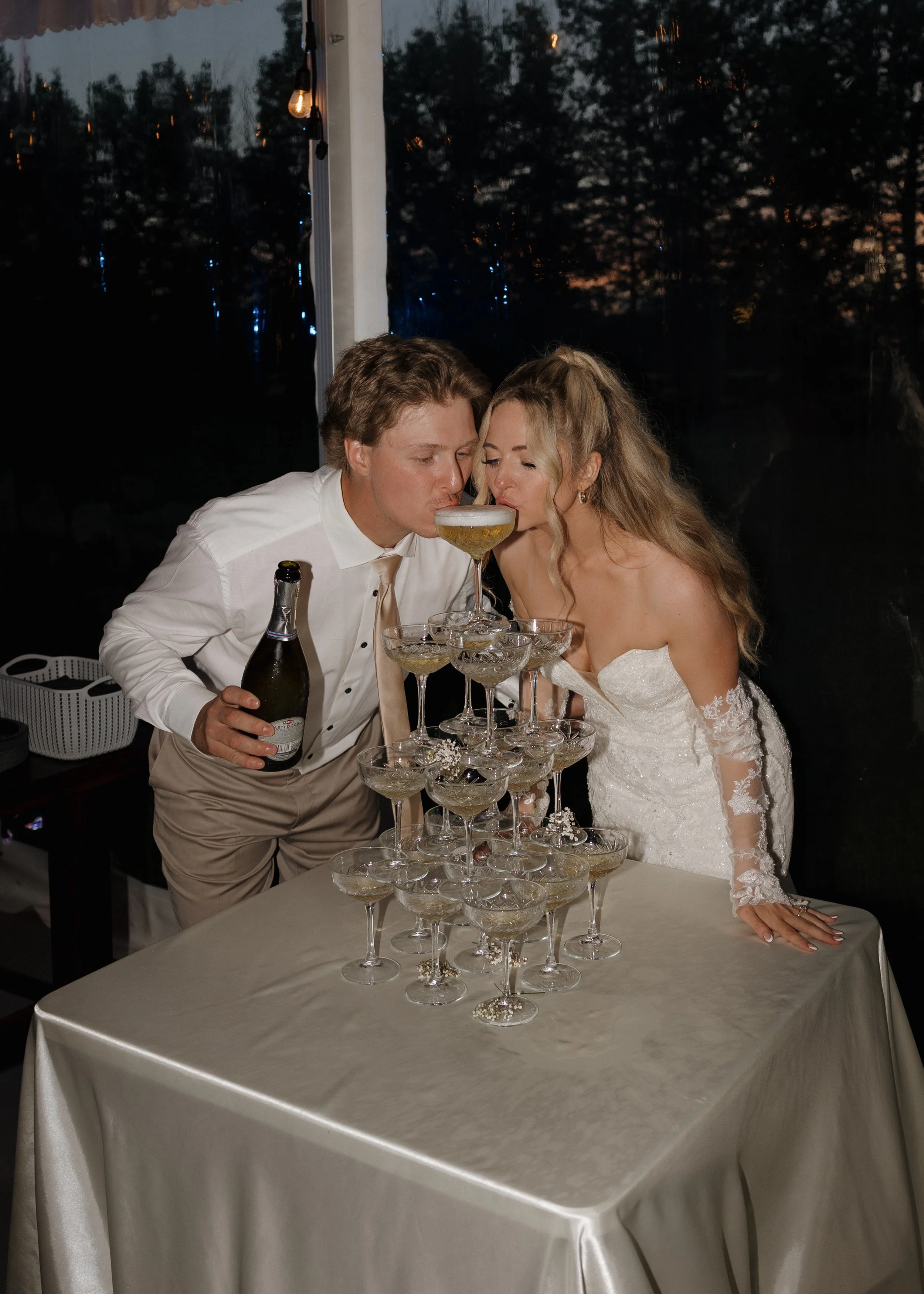 Two people, a man and a woman, at a party, sharing a toast with champagne glasses balanced on a pyramid-shaped tower of glasses on a table, with a dark outdoor background visible through a window.