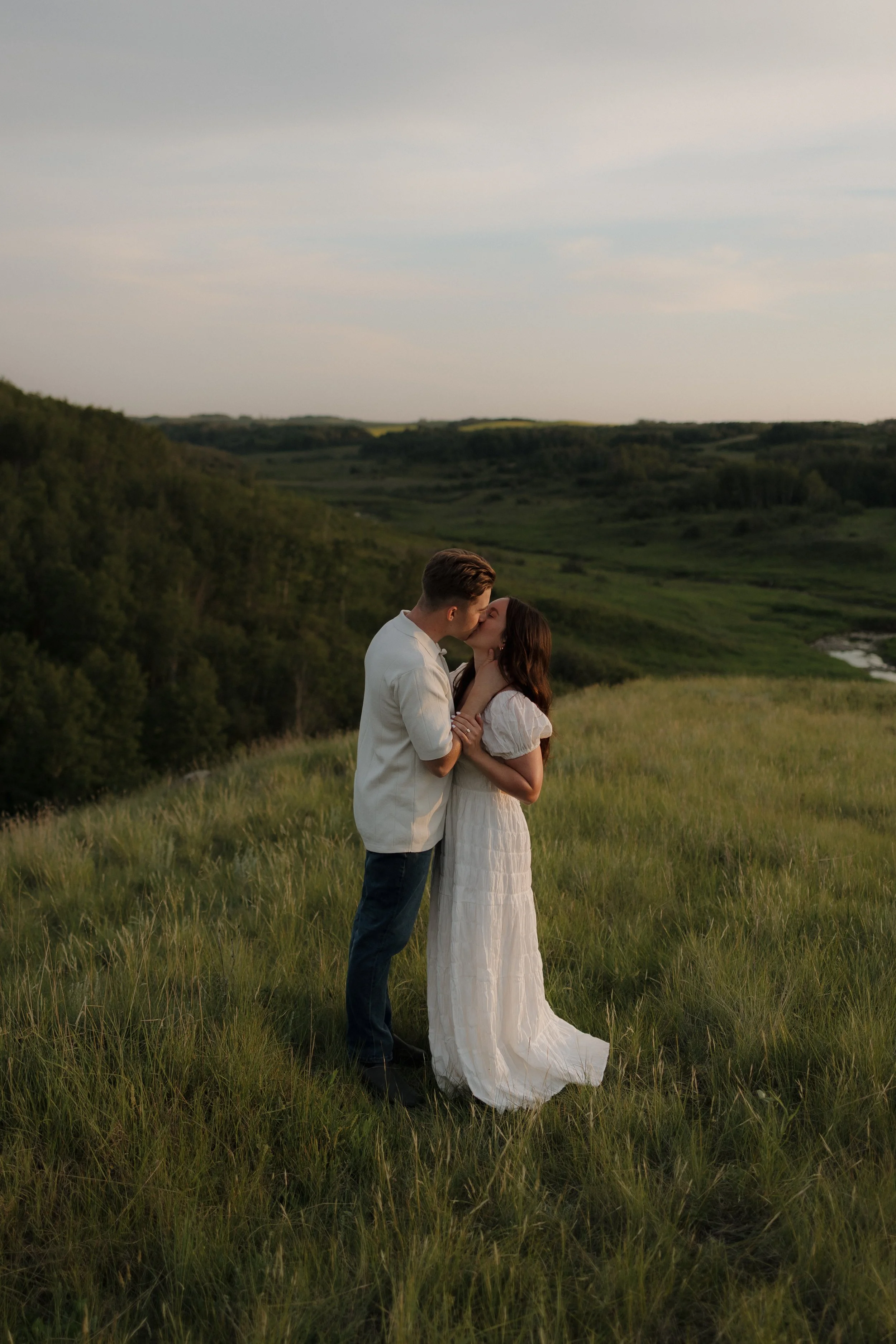 A young couple kissing in a grassy field during sunset with a scenic green landscape in the background.