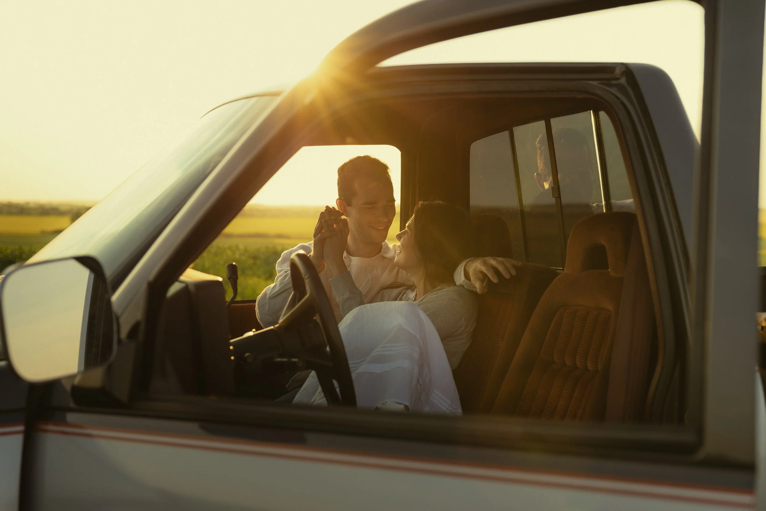 A young couple sitting in a truck holding hands while the sun shines in