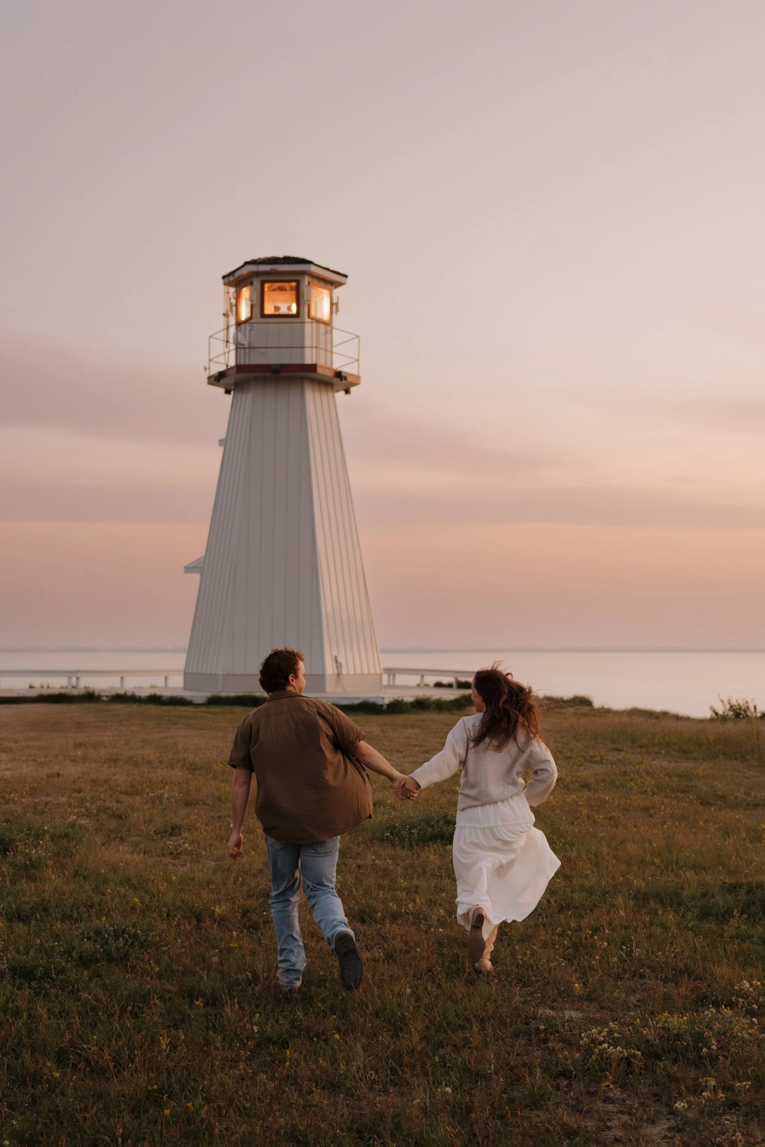 A couple holding hands and running on a grassy field towards a lighthouse during sunset.