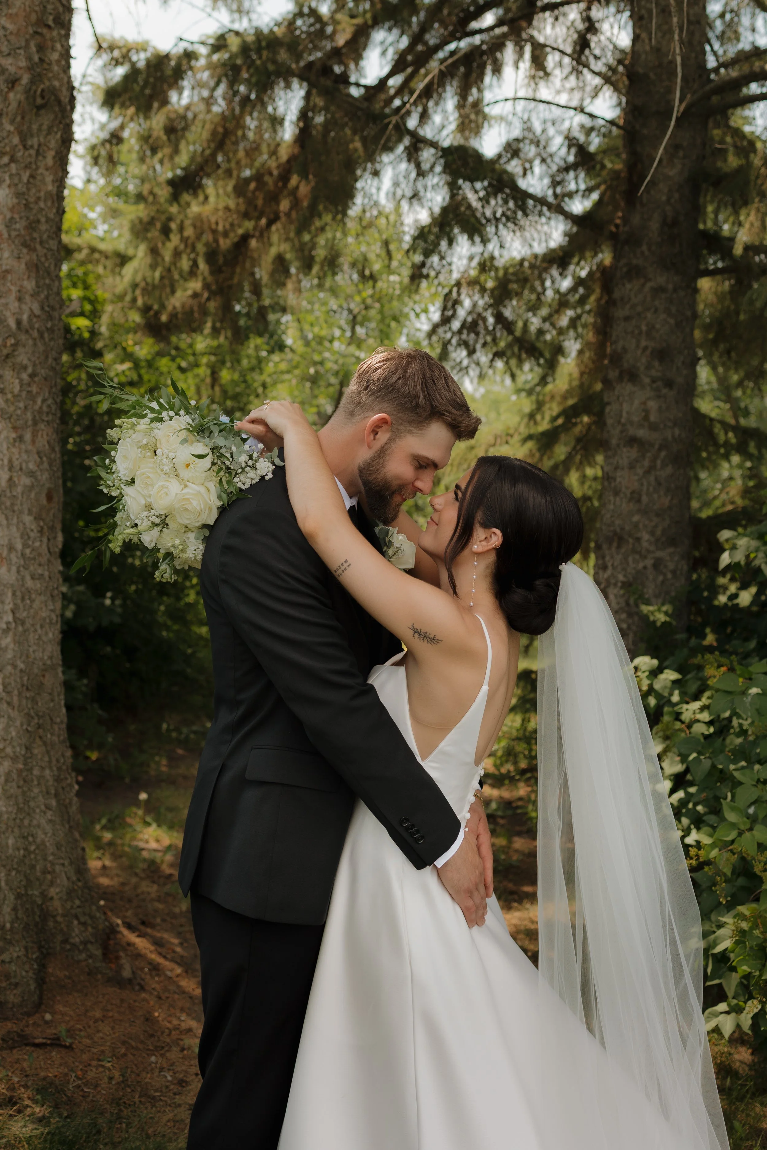 A bride and groom sharing a romantic moment outdoors during their wedding, surrounded by trees and greenery.