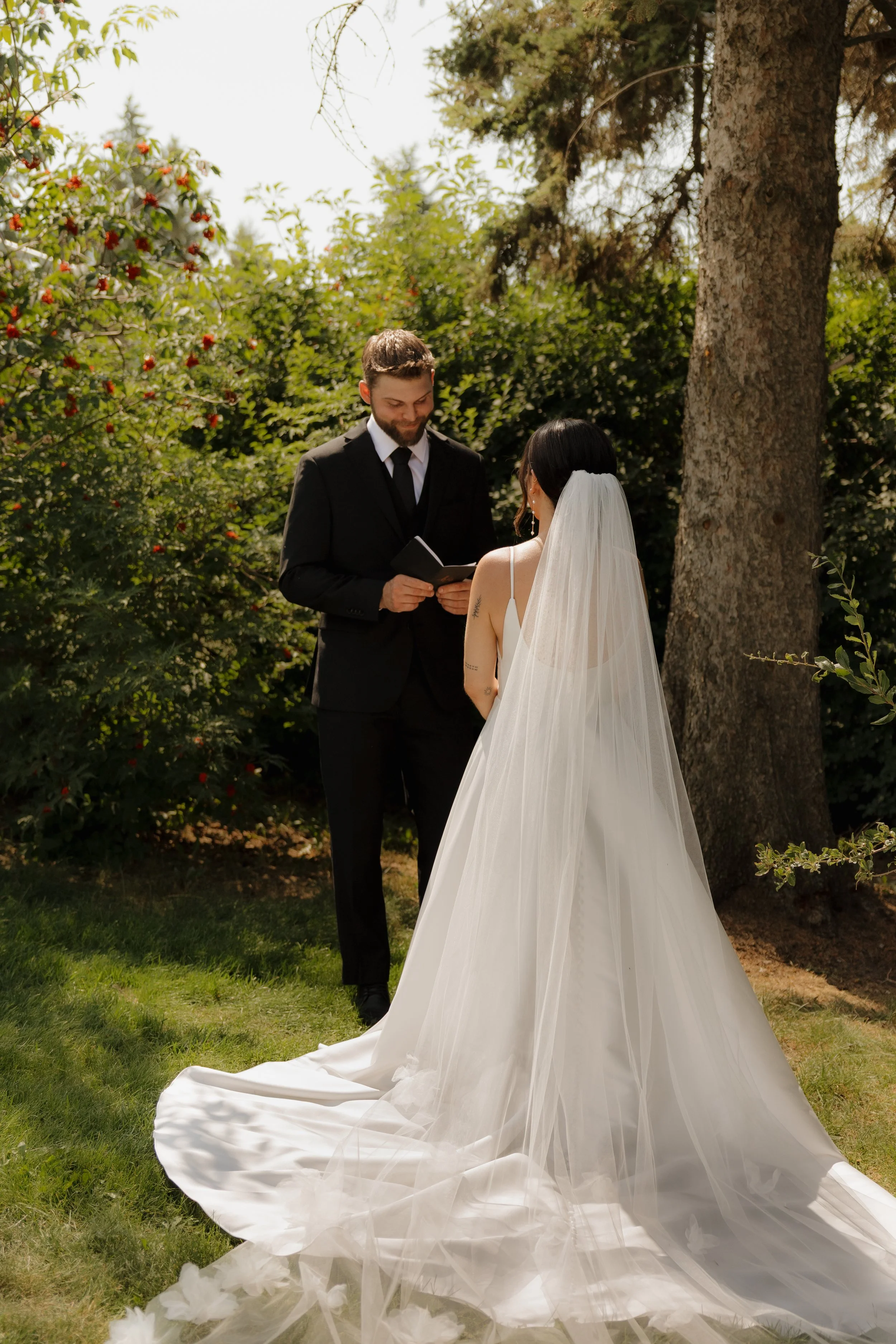 A bride and groom exchanging vows outdoors during a wedding ceremony, with trees and greenery in the background.