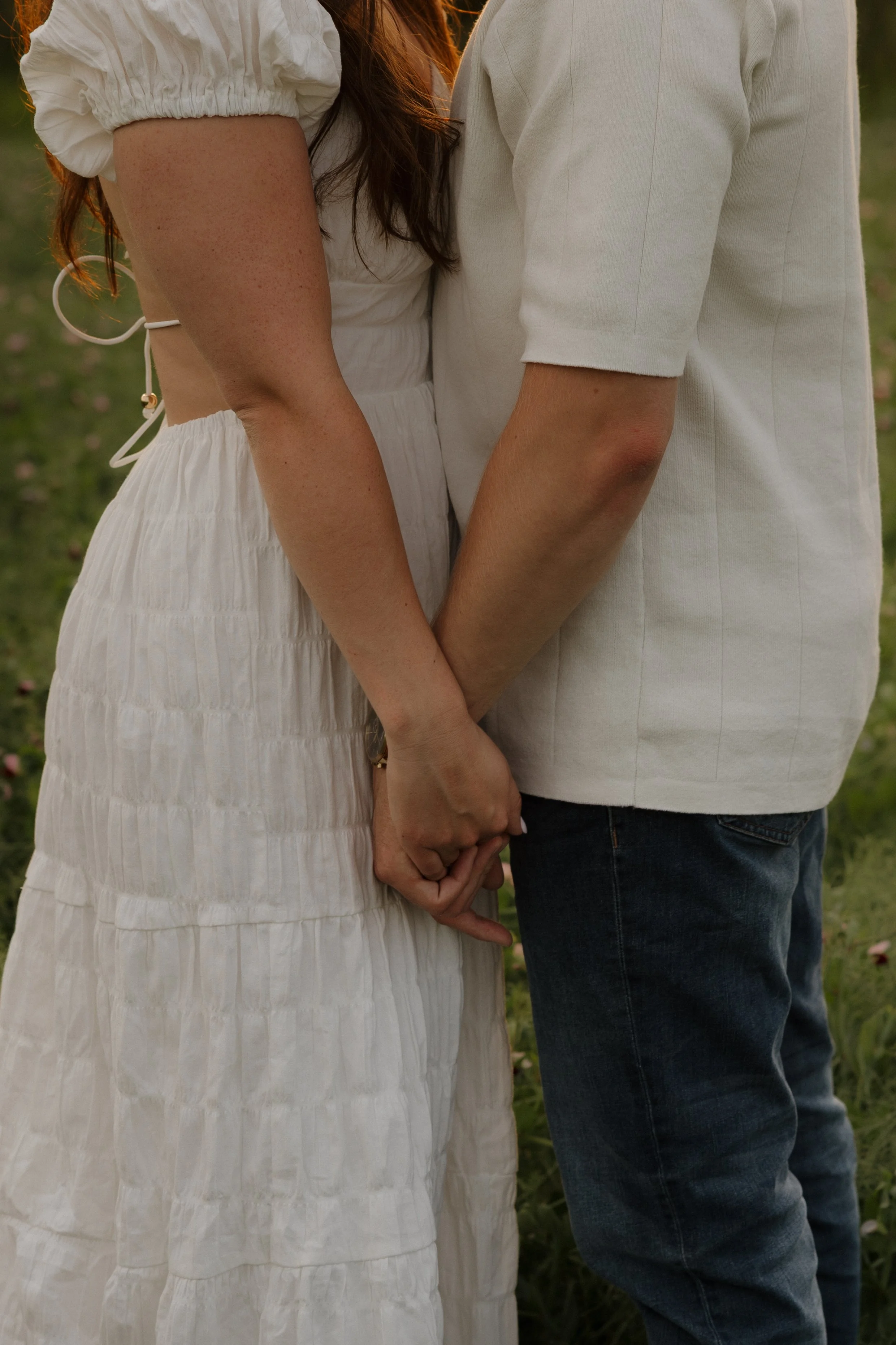 Close-up of a couple holding hands in a field, with the woman's white dress and the man's white shirt visible.