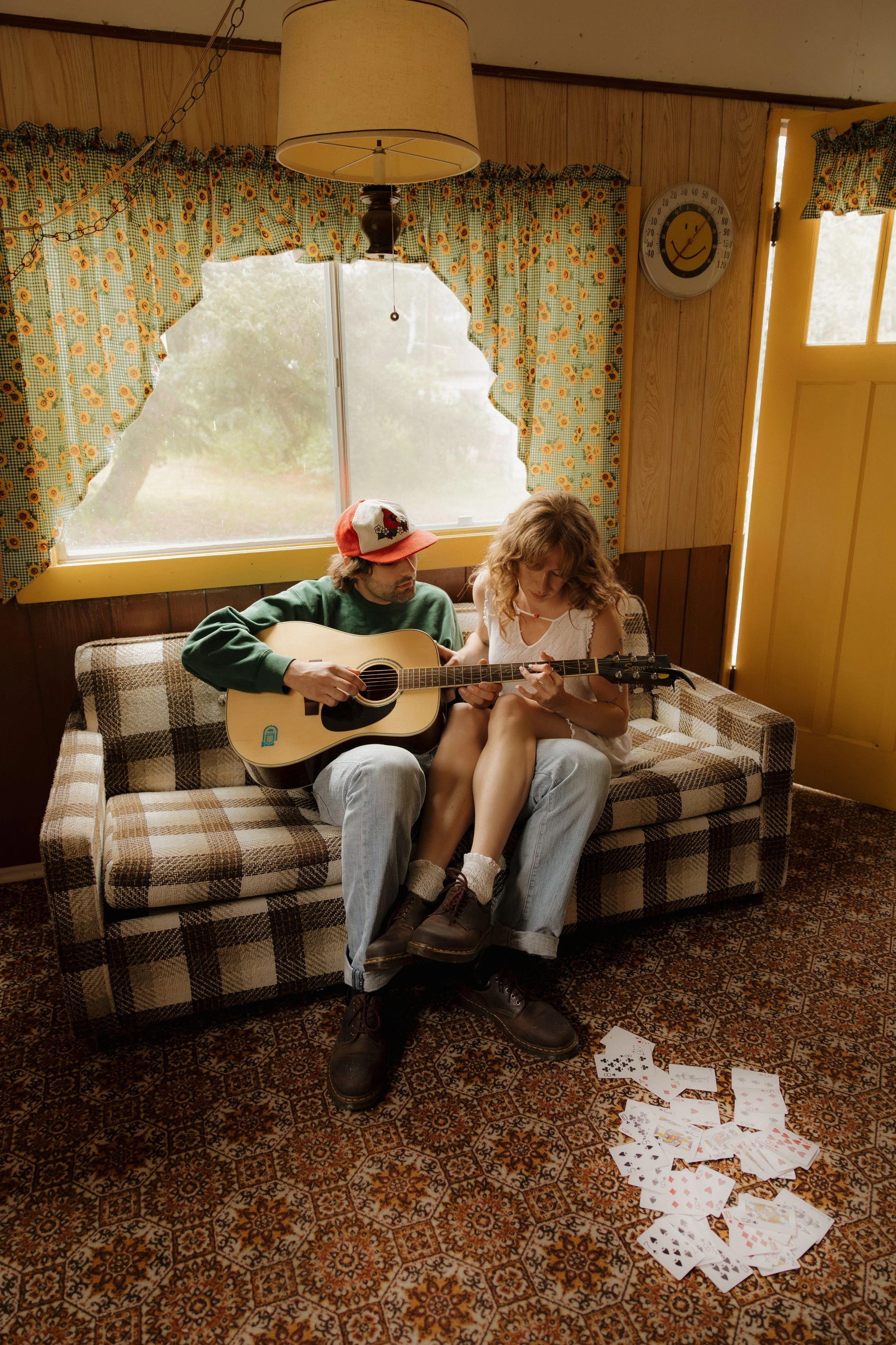 A young man and woman are sitting on a checkered vintage sofa in a cozy, rustic room. The man is playing an acoustic guitar while the woman looks at something in her hands. There are playing cards scattered on the carpet in front of them. The room ha
