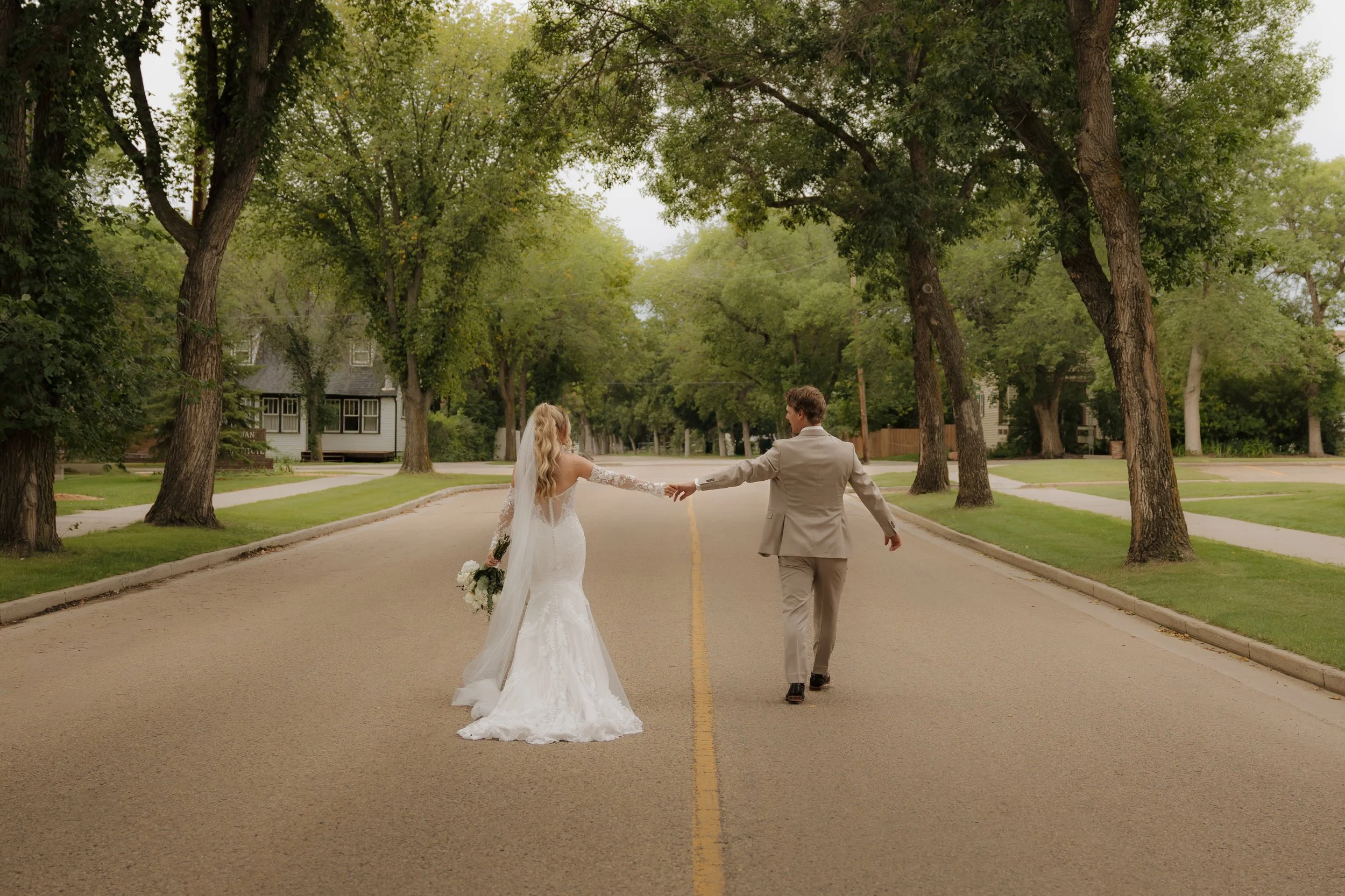 A bride and groom holding hands and walking down an empty street lined with trees and houses on a cloudy day.