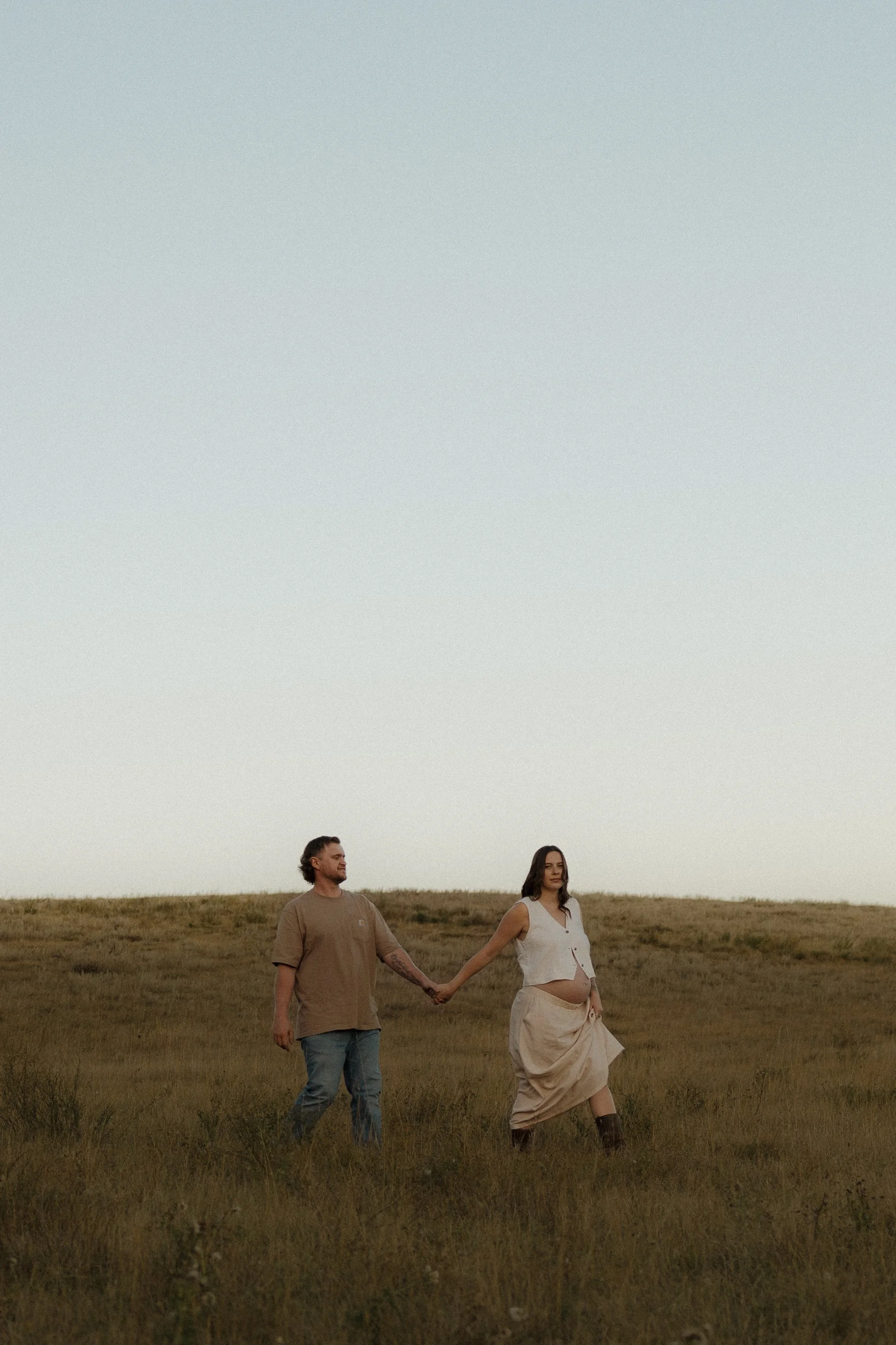 A man and pregnant woman holding hands in an open field at sunset.