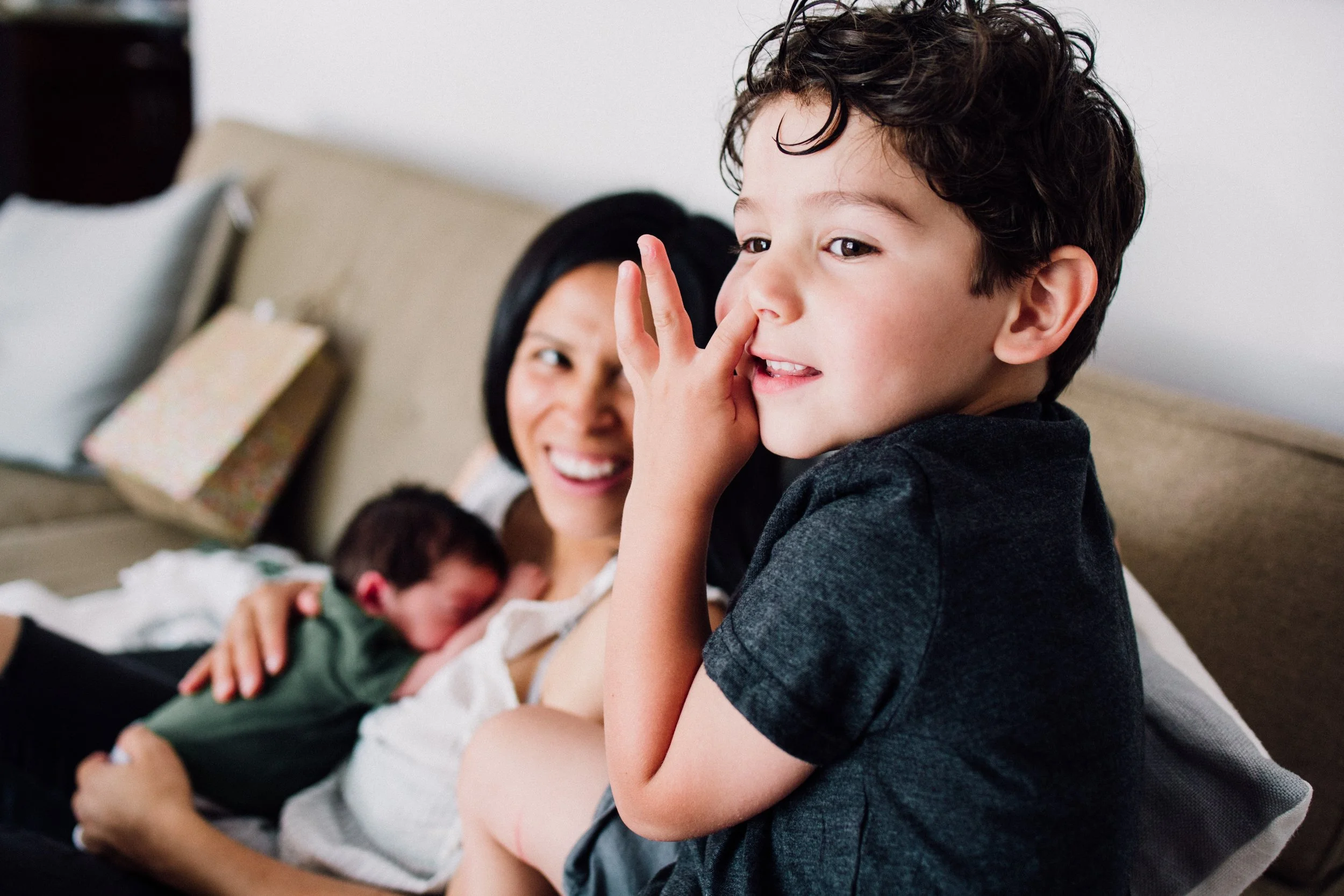 A young boy with curly dark hair sitting on a couch, with a woman and a newborn baby in the background. The boy is touching his nose with his finger, and the woman is smiling.