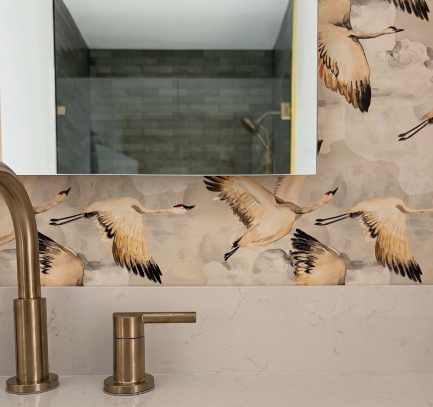 Bathroom with wallpaper featuring flying cranes, a golden faucet, and a mirror reflecting a shower area with gray tiles.