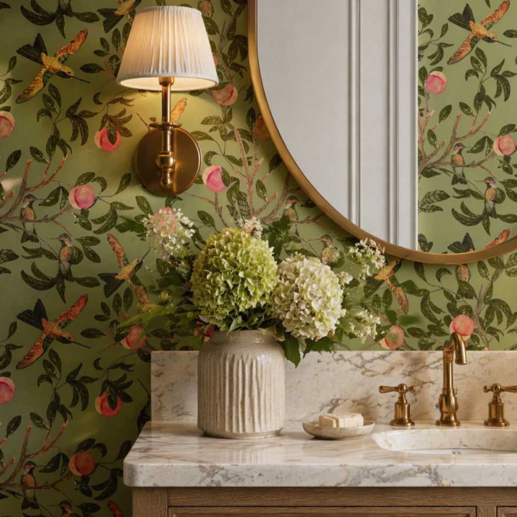 A bathroom vanity with a pink cabinet and a white marble countertop. On the countertop is a glass vase with white and light purple flowers. Above the vanity is a round mirror with a silver frame. To the left of the mirror is a wall-mounted light fixture with a white shade. The wall behind has green wallpaper with a floral pattern of branches, leaves, pink flowers, and hummingbirds.