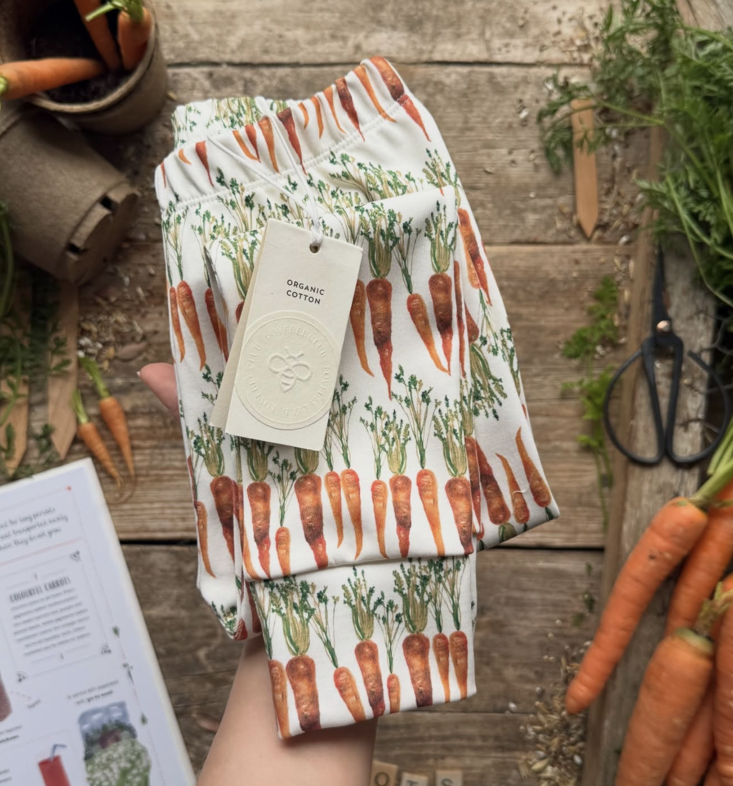 A person holding a pair of pants with a carrot print, a tag indicating they are made of organic cotton, in a setting with fresh carrots, scissors, and gardening supplies on a wooden surface.