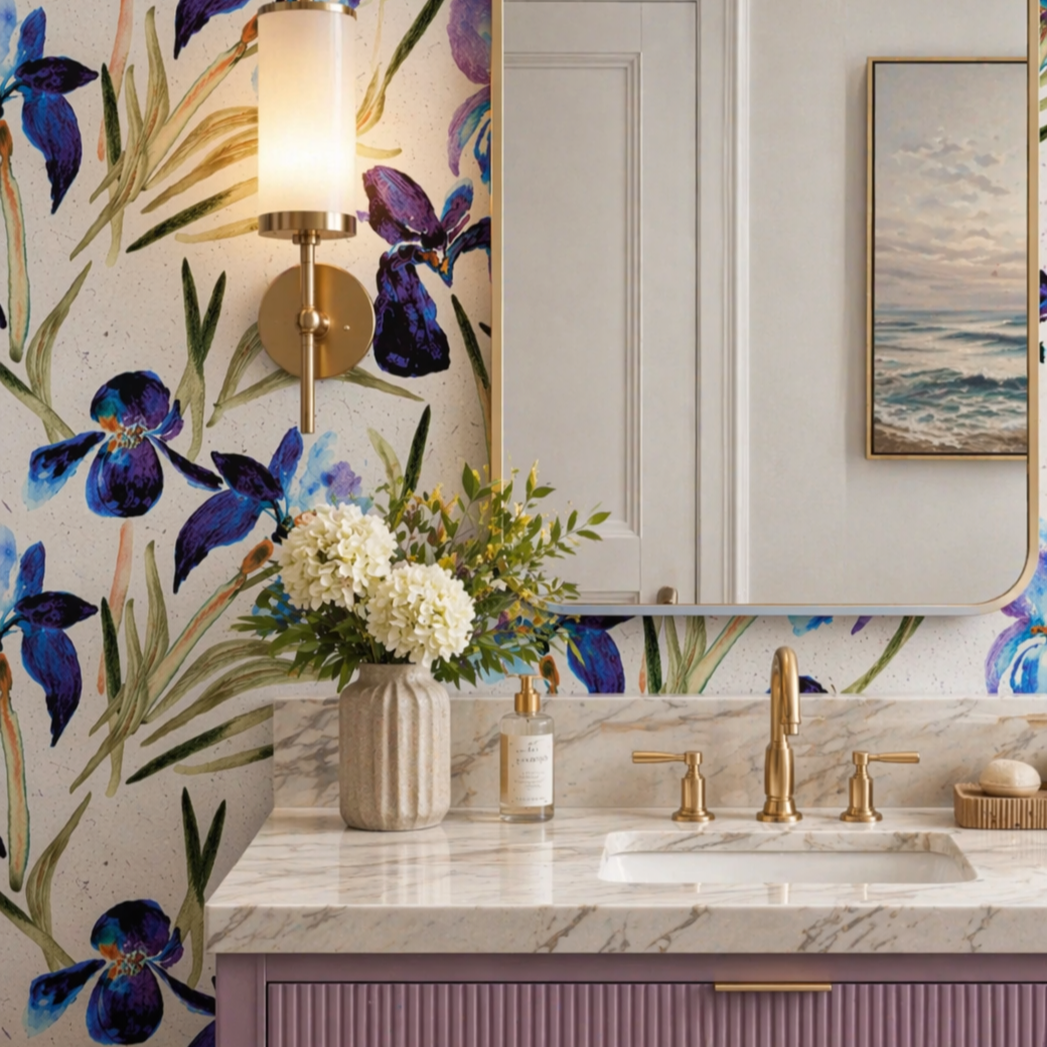 A bathroom vanity with a purple cabinet and a white marble countertop. A clear glass vase with beige and white flowers is on the counter. Behind, a round mirror frames a white door, and a wall sconce with a white shade is mounted on a floral wallpaper featuring purple and blue flowers with green leaves.