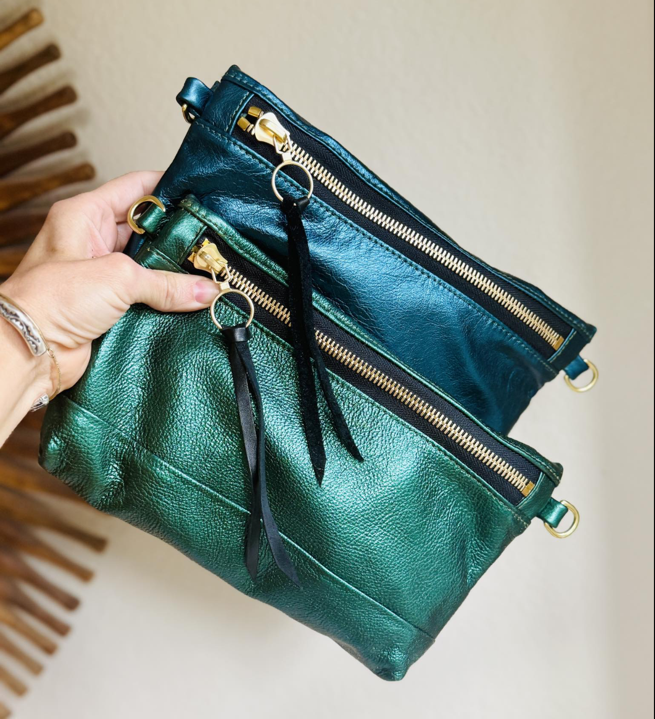 A hand holding two metallic green and blue leather zippered pouches with gold zippers and black ribbons attached, against a beige wall and wooden decor.