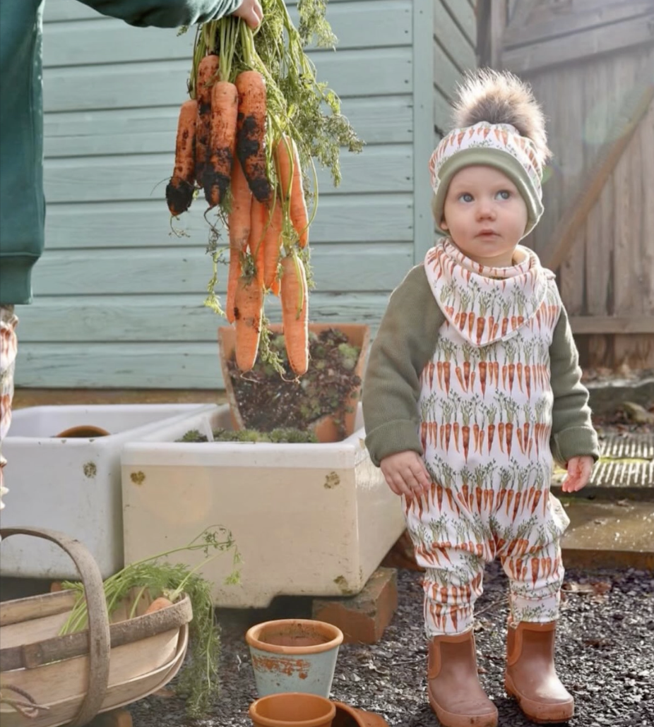 A young child dressed in carrot-themed clothing standing outdoors near a garden with carrots hanging from a hand, some potted plants, and garden containers.