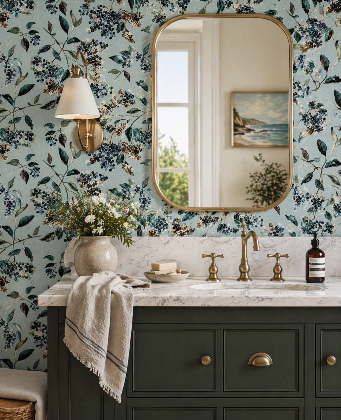A bathroom vanity with a green cabinet, white marble countertop, a glass vase with white flowers, and a large oval mirror. The wall has floral wallpaper with dark berries and leaves, and there is a wall sconce with a white shade.