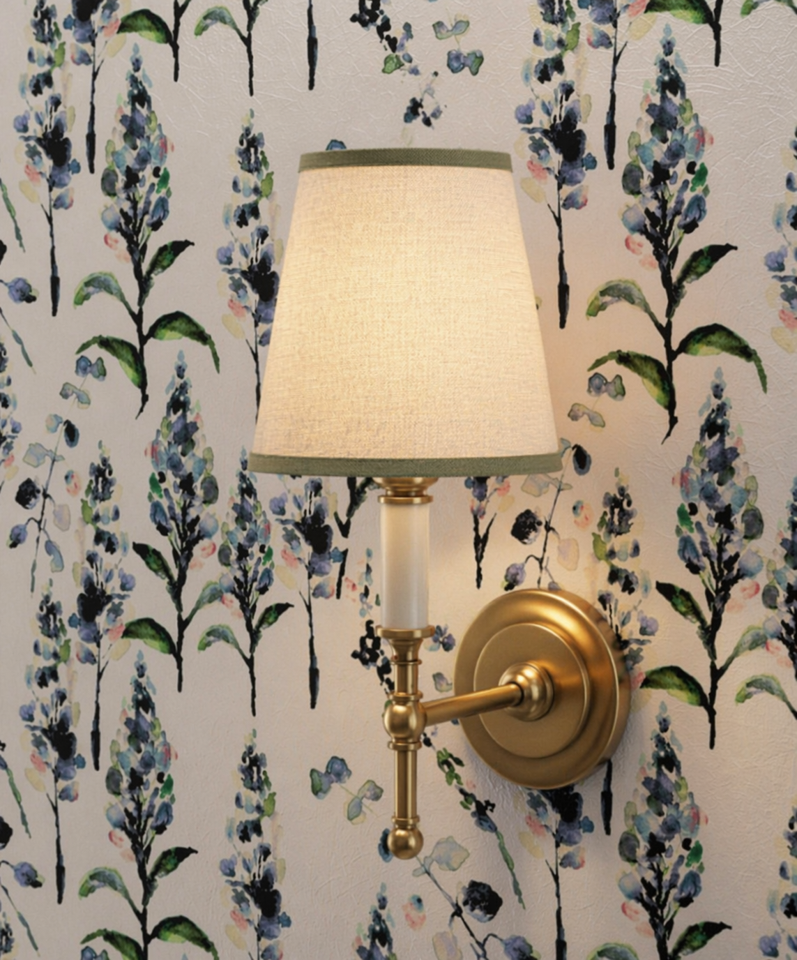 Wall-mounted gold sconce with white lampshade in front of wallpaper with floral pattern of blue, purple, and green flowers and black stems.