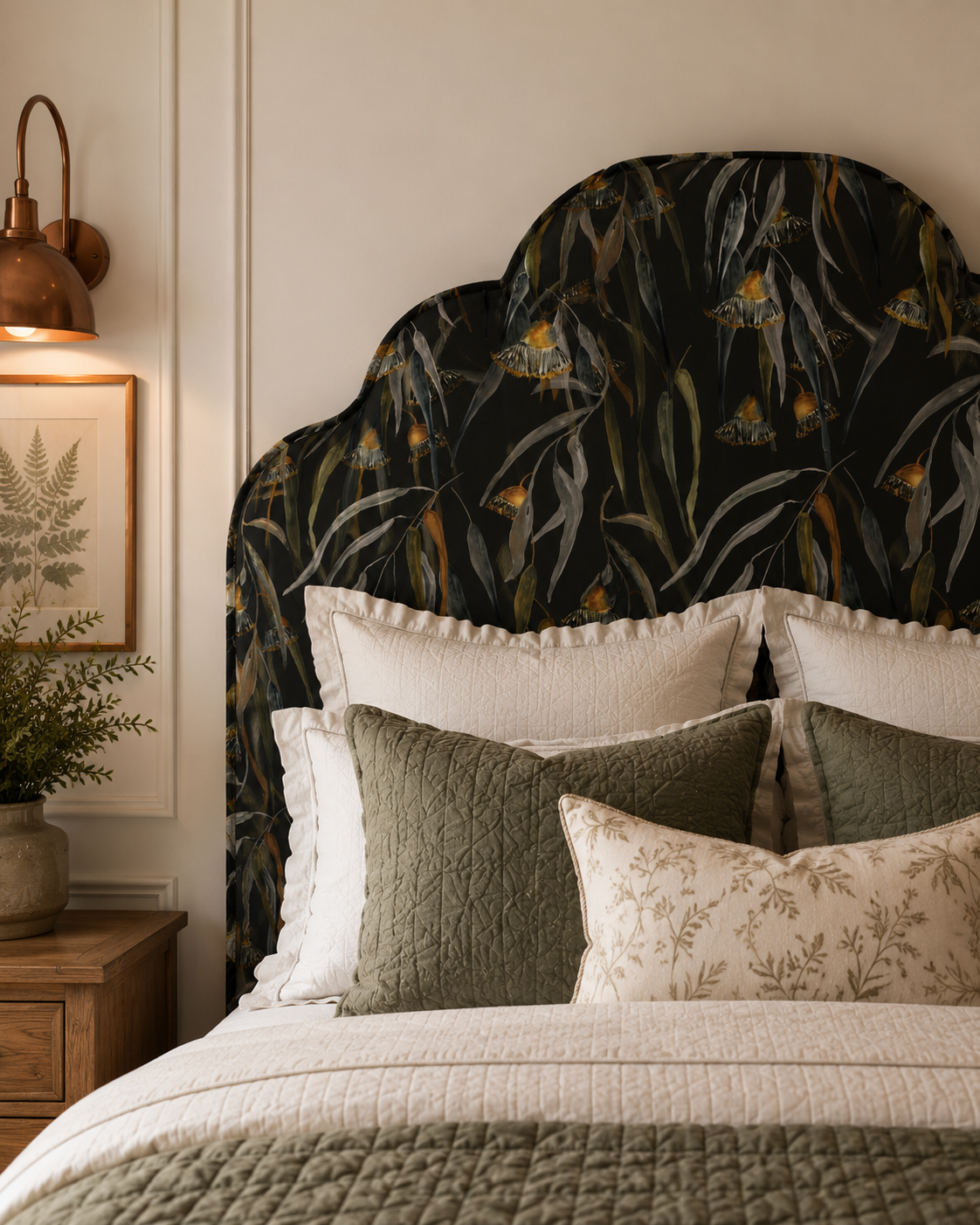 A cozy bedroom with dark green walls, a botanical wallpaper feature wall, and a green upholstered bed with pillows in teal, terracotta, and gold. There are two pendant lights hanging from the ceiling, a bedside table with a lamp and candles on each side, and a brown tufted bench at the foot of the bed with books and a gray throw blanket. A rug covers the wooden floor.