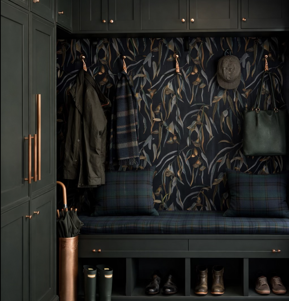 Entryway with dark green storage cabinet, hooks with jackets, scarf, and tote bag, a hat, plaid bench with pillows, and shoes underneath, against a dark botanical wallpaper.