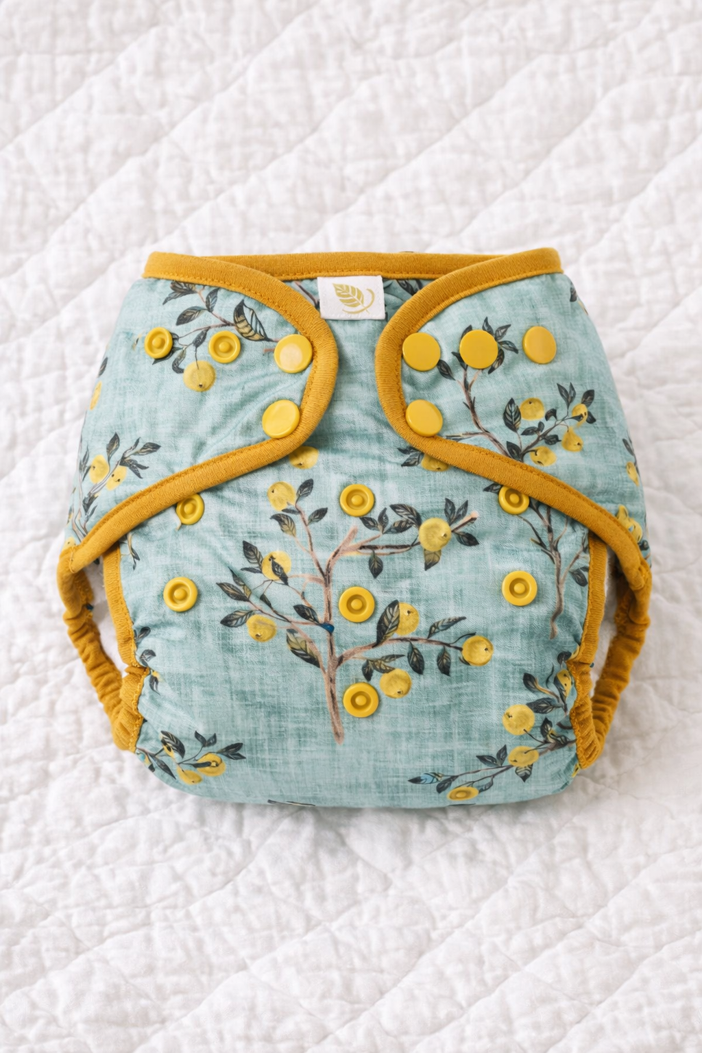 A cloth diaper with a yellow trim and snap closures on a white quilted surface, featuring a pattern of yellow berries and dark green leaves on a light green background.