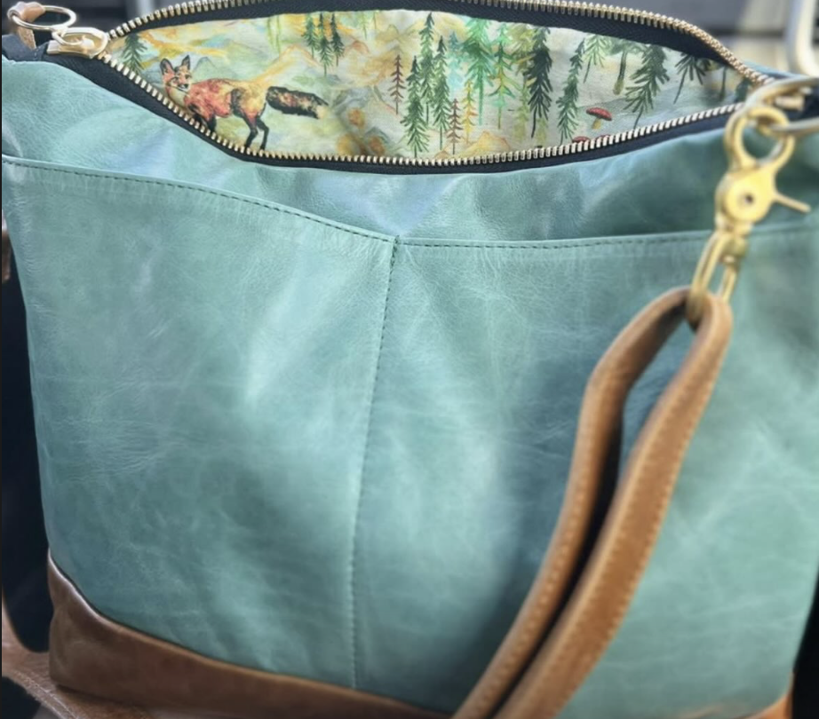 Close-up of a teal handbag with tan handles, unzipped to show a woodland animals and trees pattern inside.