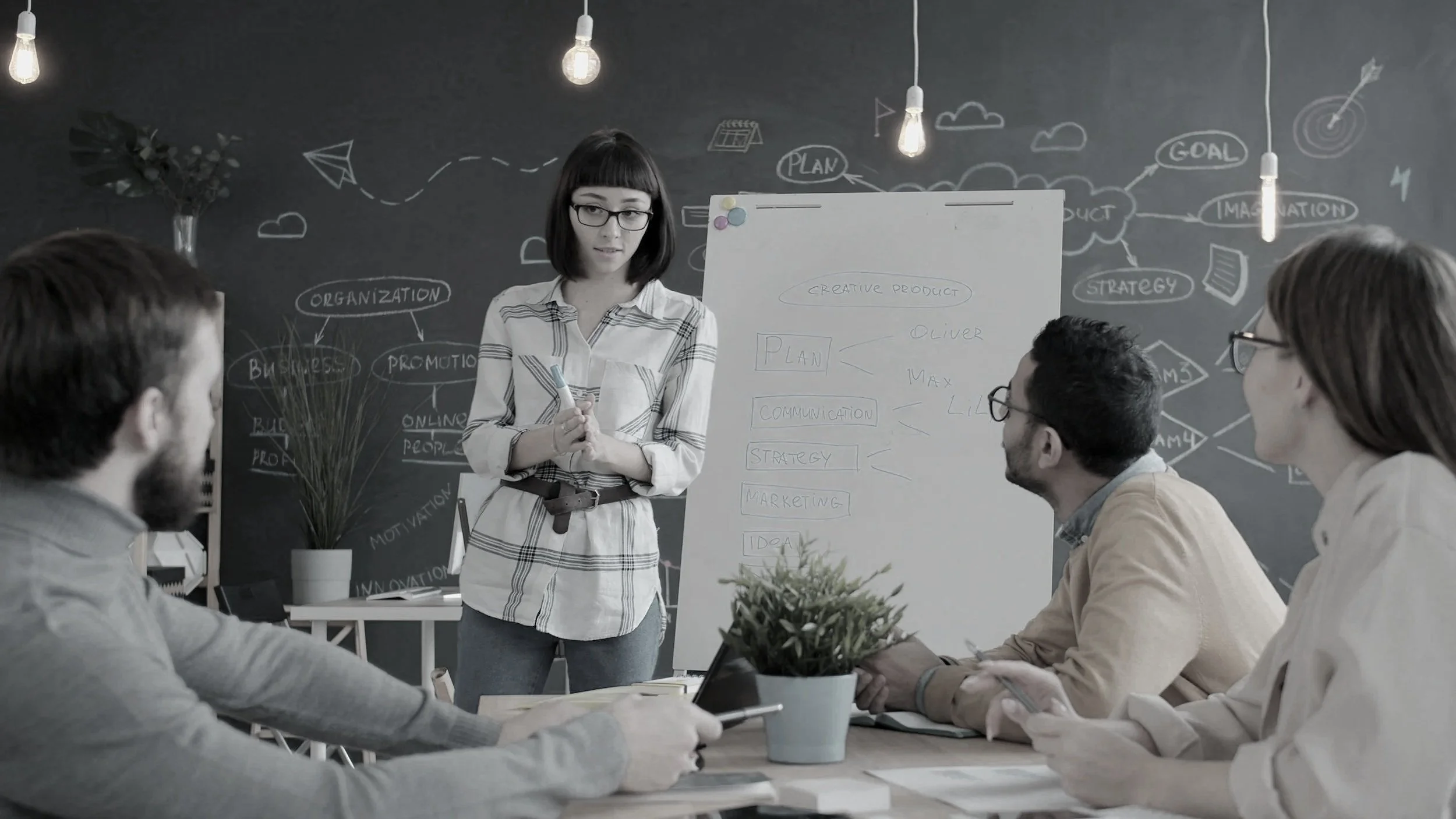A woman presenting to a group of colleagues in a meeting room, with diagrams and notes on a whiteboard and chalkboard behind her.