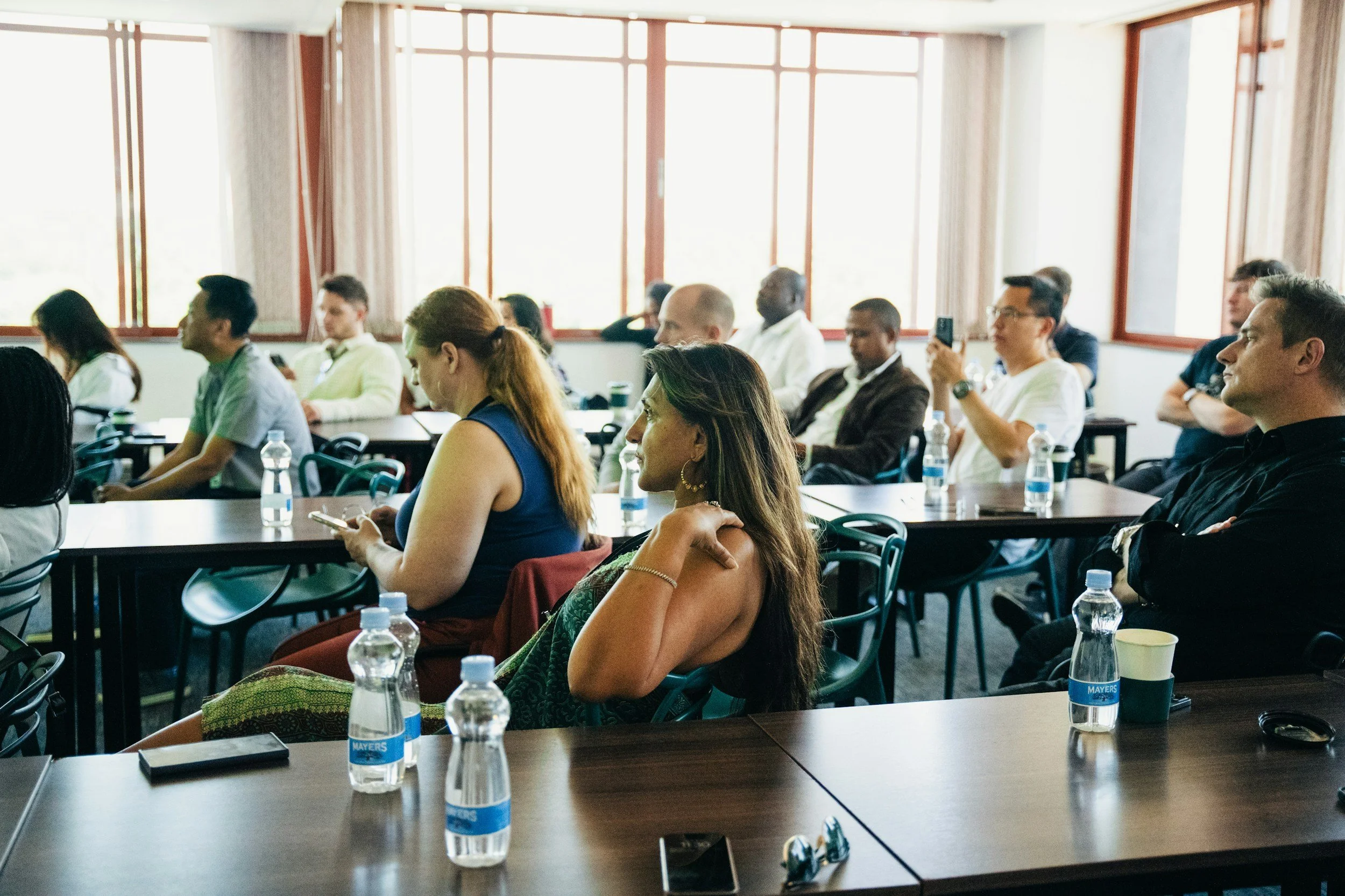 Attendees seated at a conference table listening to a presentation in a bright room with large windows.