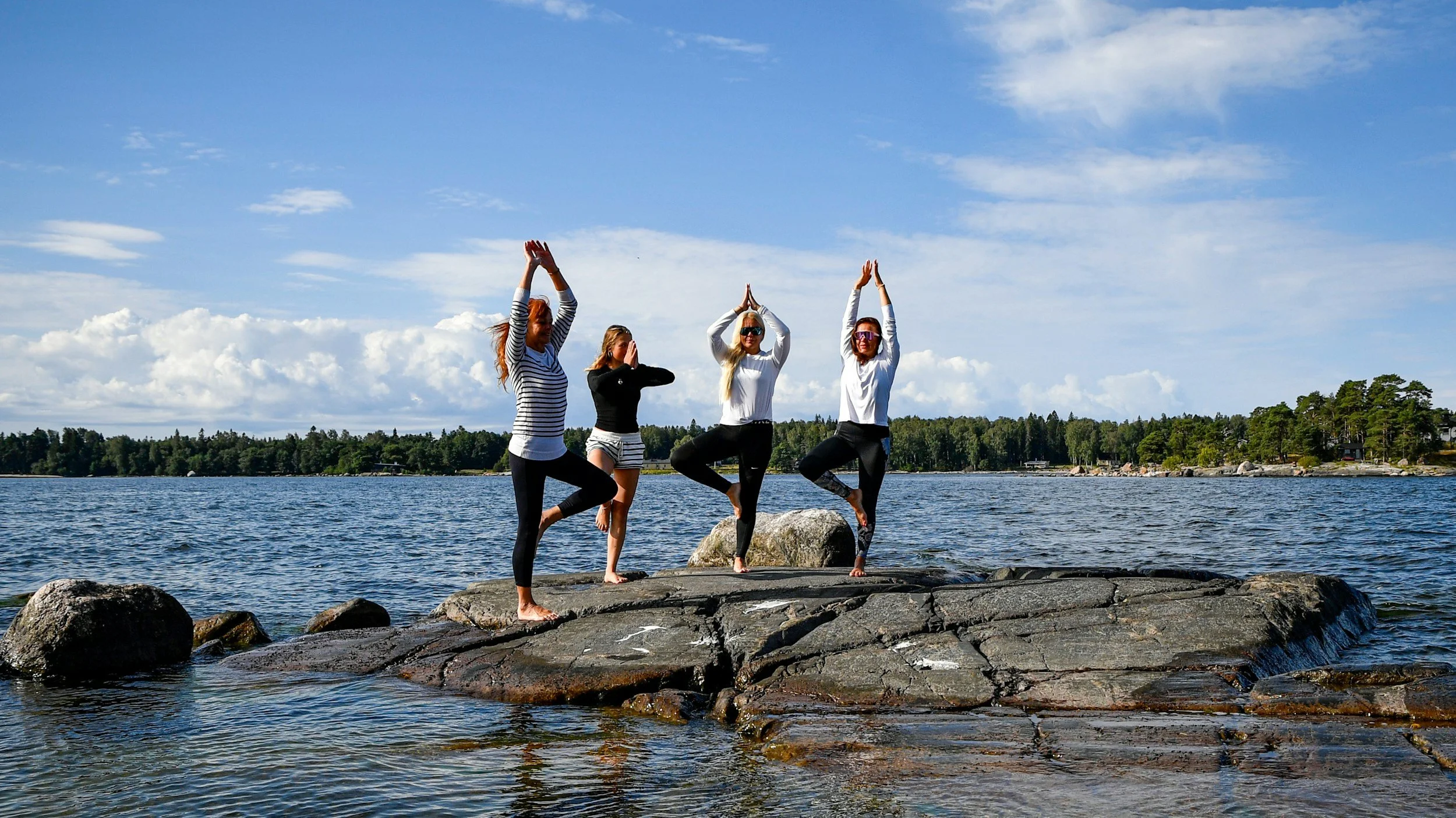 Four women practicing yoga on rocks by the water on a sunny day.