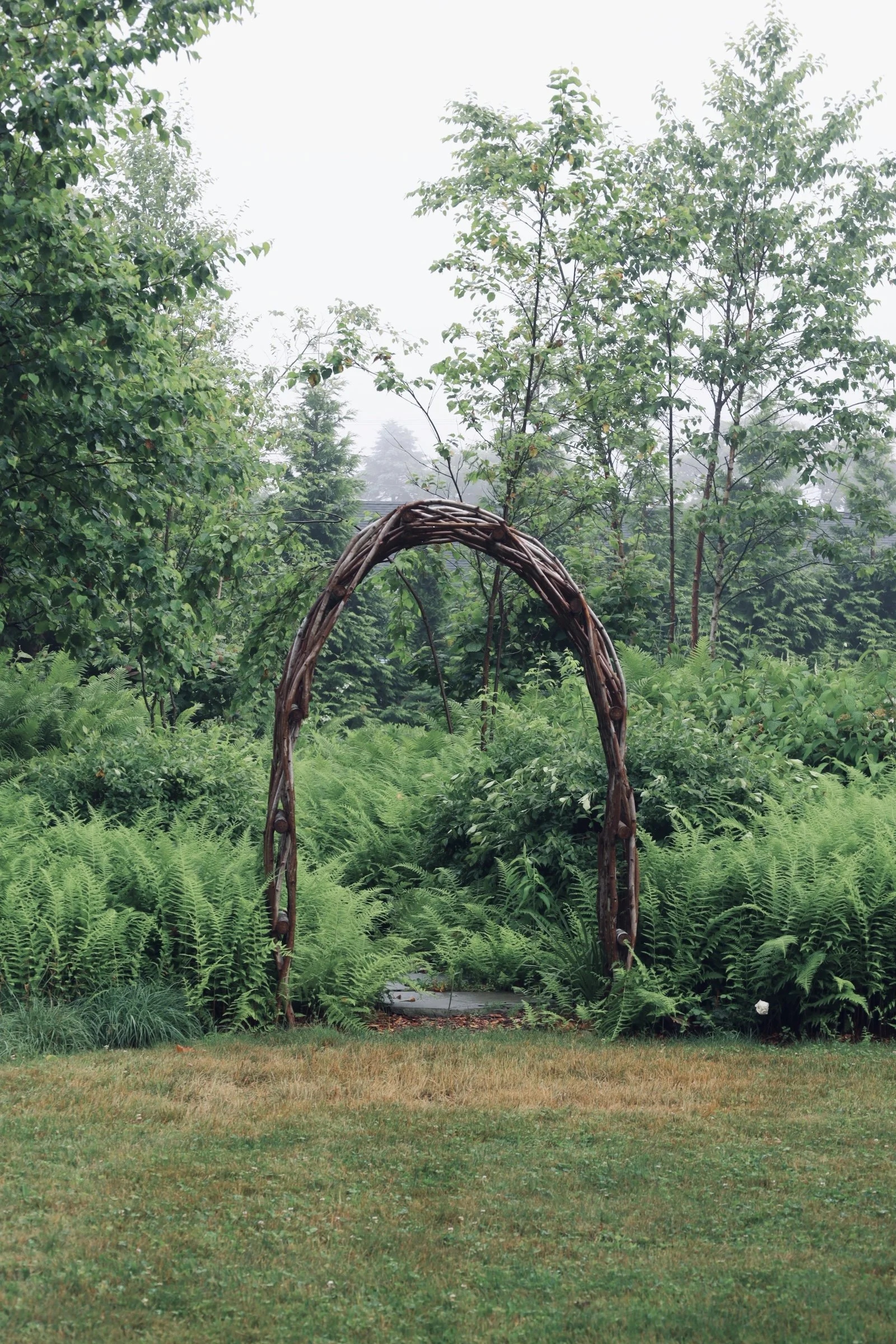 A garden scene with a wooden archway surrounded by lush green plants and trees.