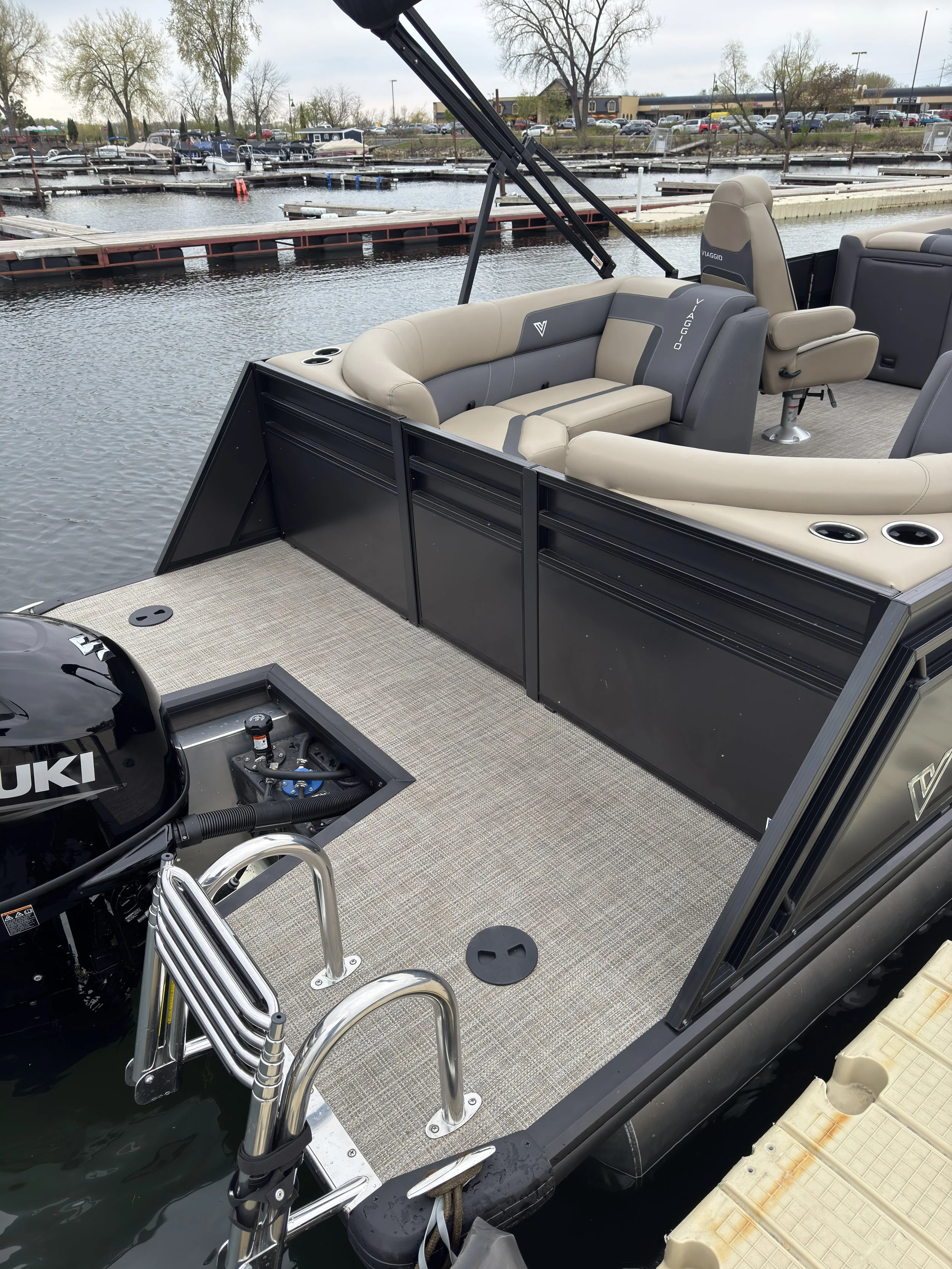 Image shows the interior of a pontoon boat docked at a marina. The boat has beige and gray cushioned seats, cup holders, a metal ladder, and a black outboard motor. The marina with docks and boats, trees, and buildings can be seen in the background.