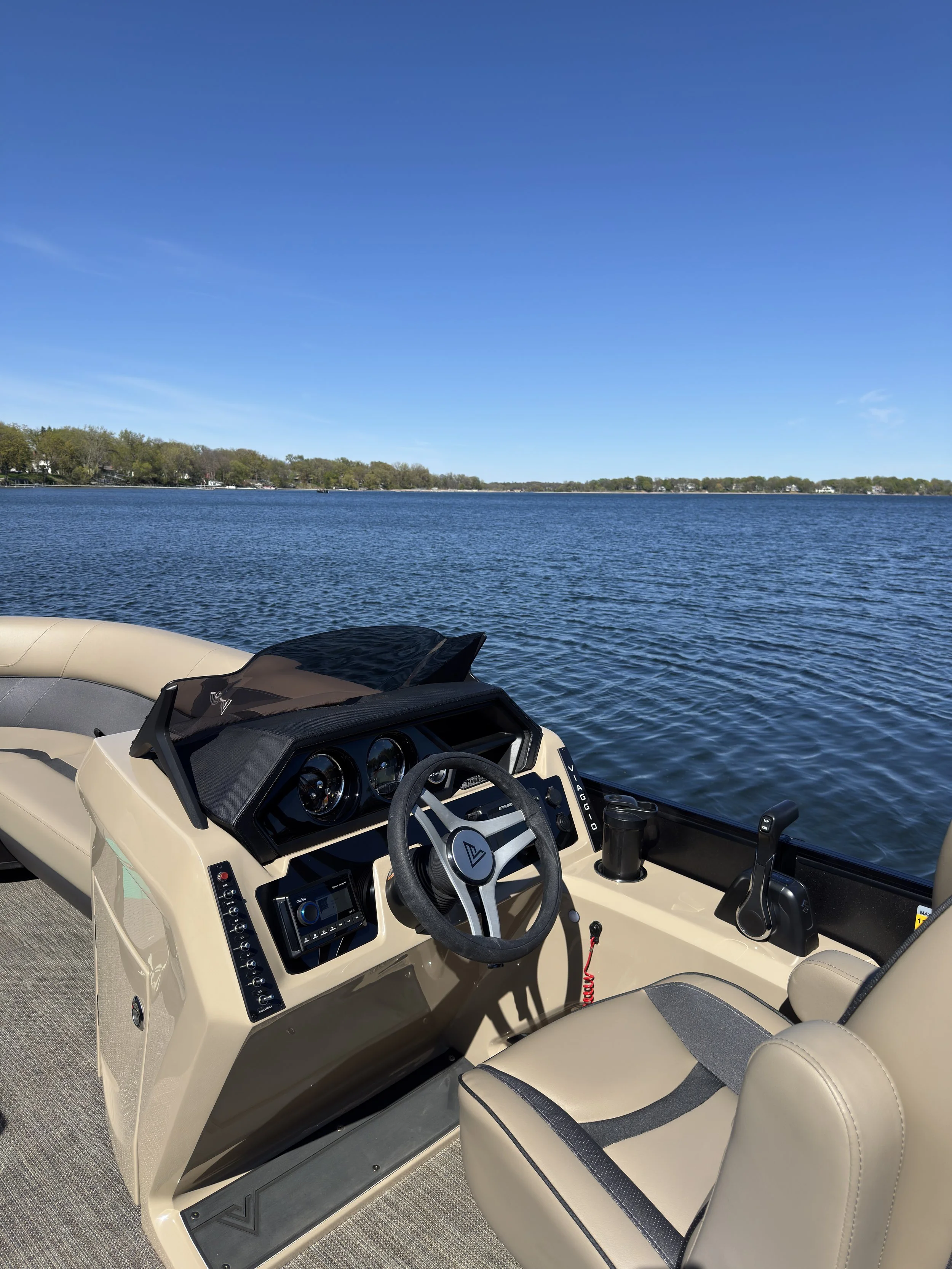 The image shows the interior of a boat with a beige and black dashboard, steering wheel, and seats. The boat is on a body of water with a treeline in the background under a clear blue sky.