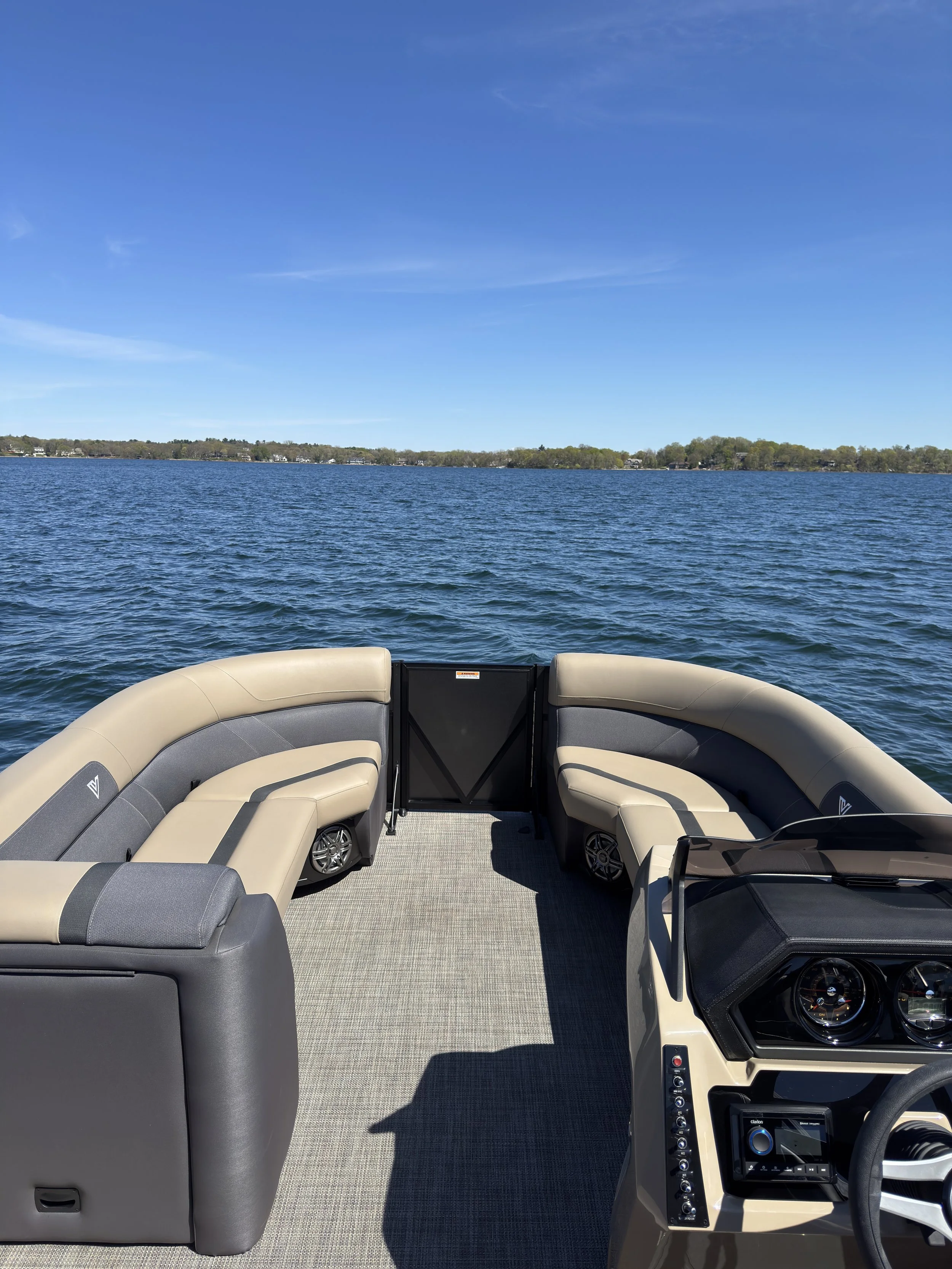 View from the back of a boat showing beige and gray cushioned seating, a steering wheel, and a dashboard with controls, overlooking a large body of water with a treeline in the distance under a clear blue sky.
