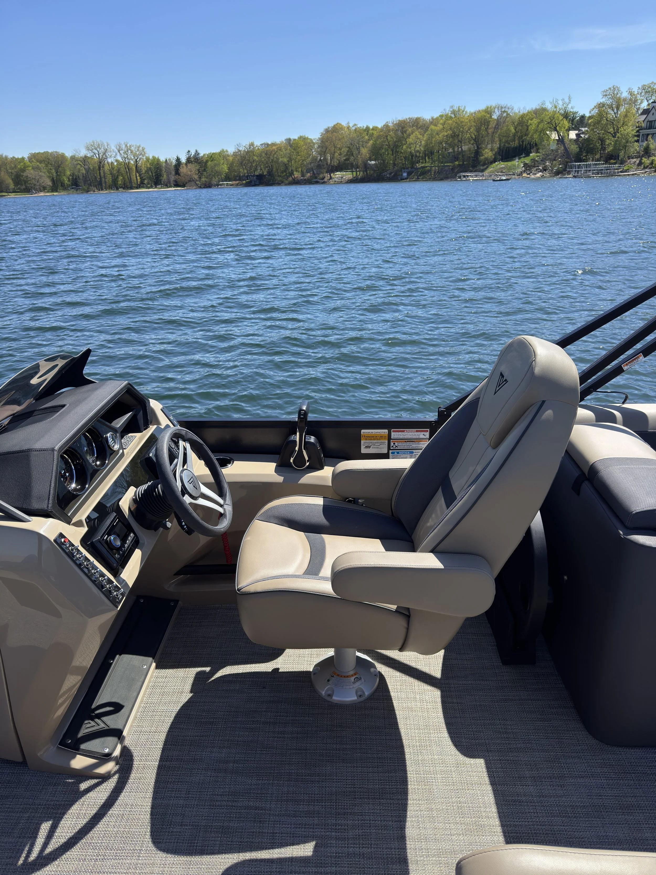View of the steering wheel and dashboard of a boat with beige seats and a scenic lake with green trees and houses in the background.