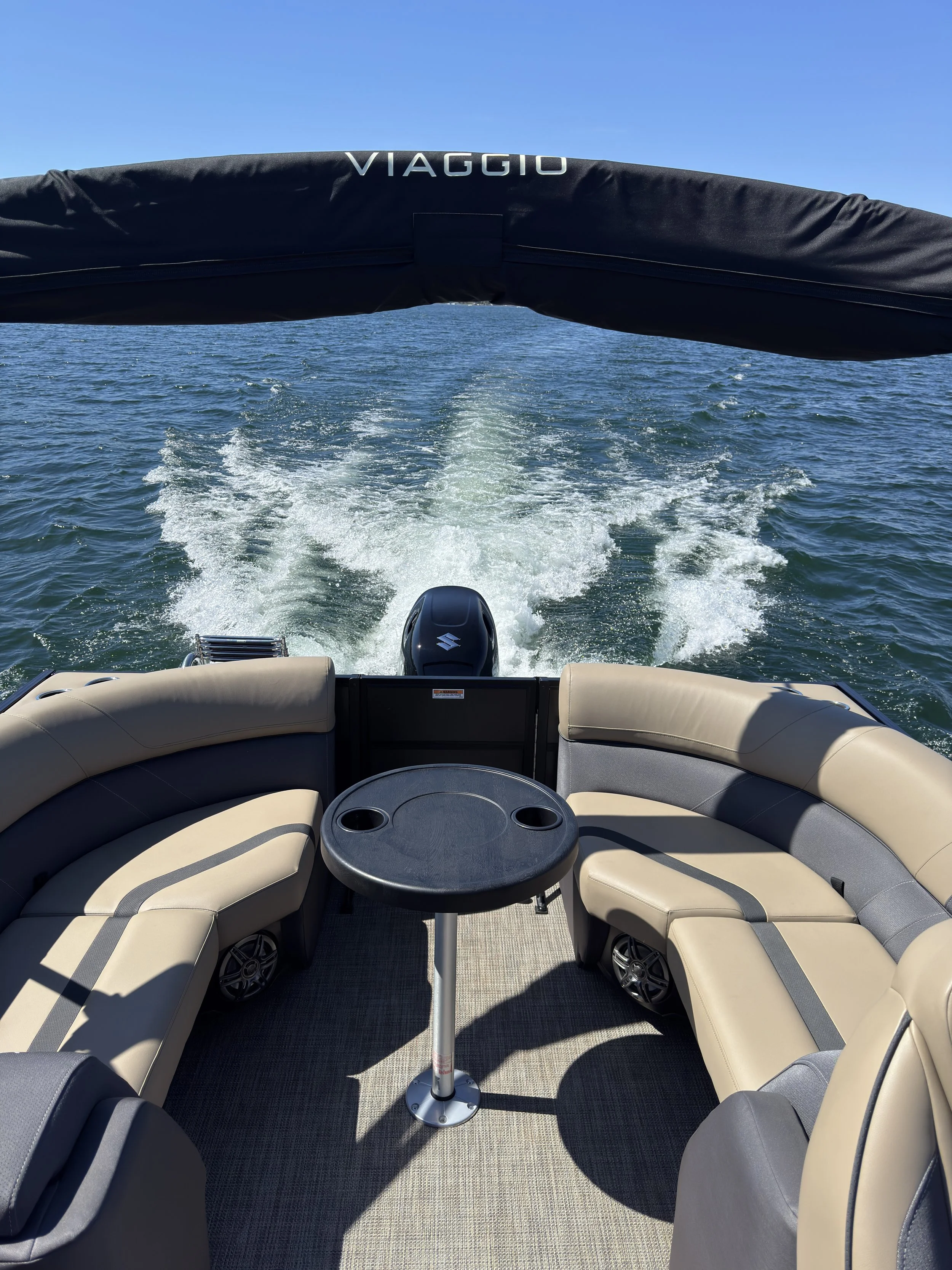 View of the back of a boat with beige and gray cushioned seating, a small round table, and a Suzuki outboard motor, traveling on a body of water on a sunny day, leaving a wake behind.