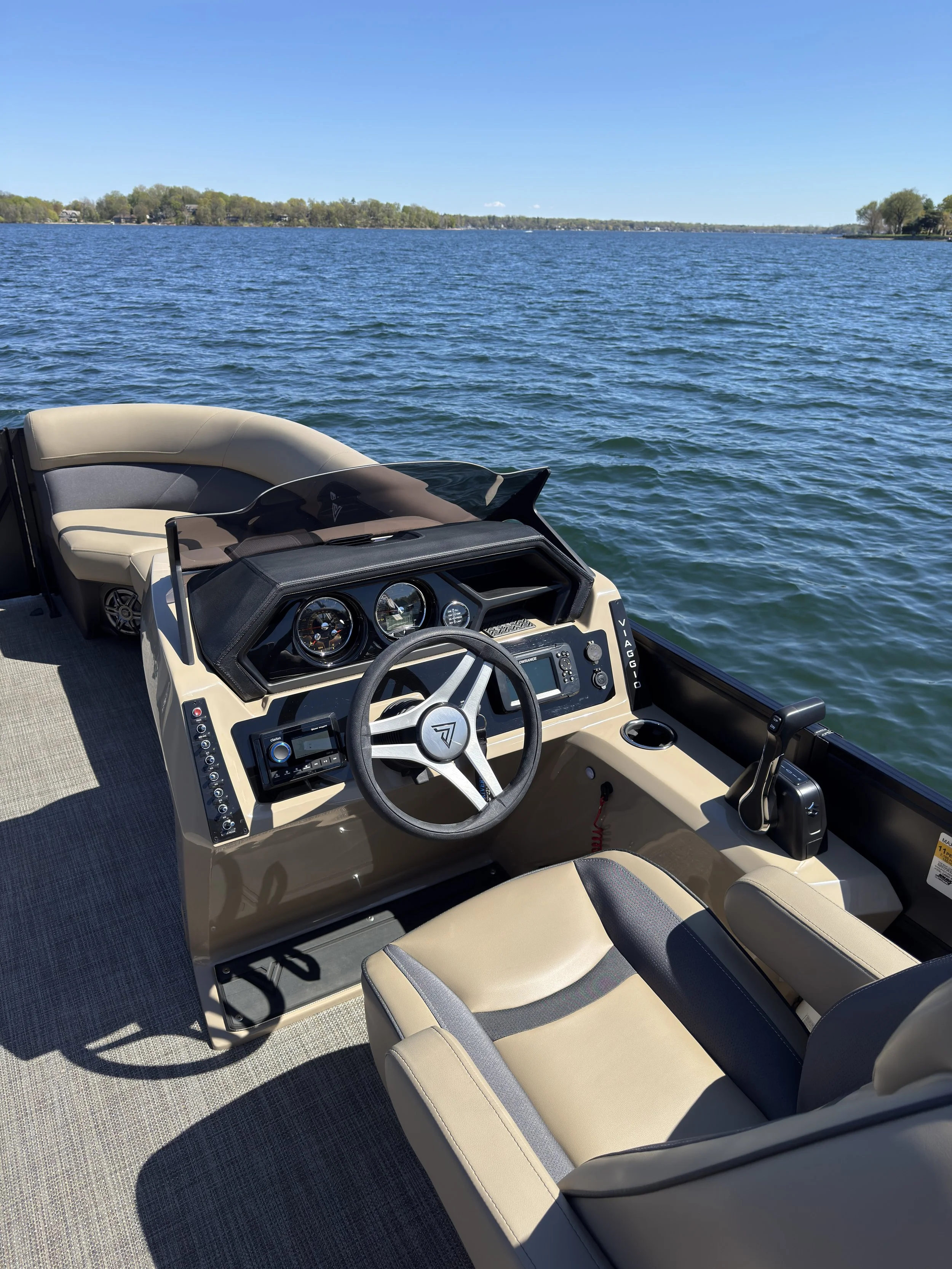 View of boat's steering console with a beige cushioned seat, steering wheel, and various gauges, overlooking a body of water with a distant shoreline and clear blue sky.