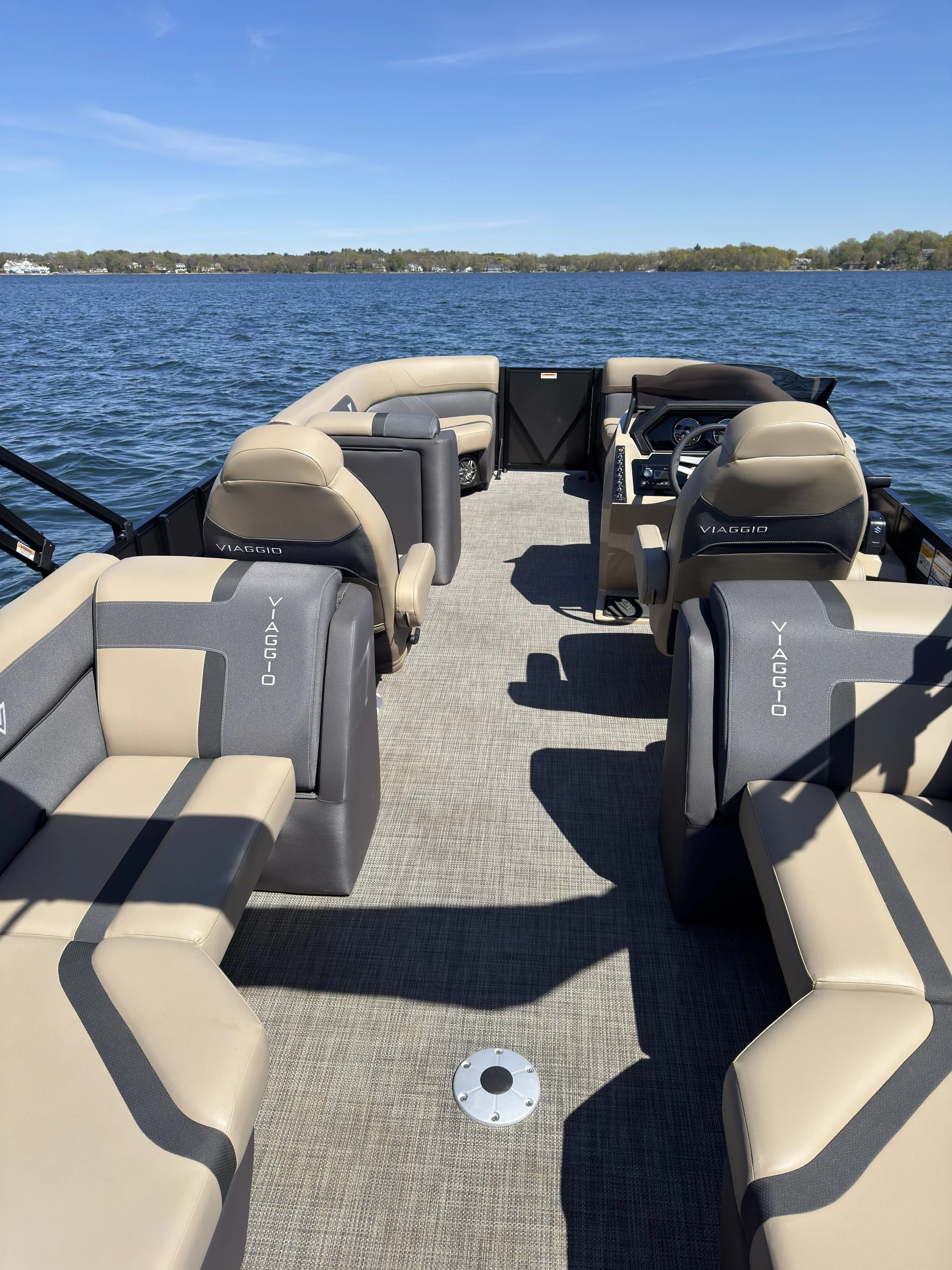 View of a boat's deck with beige and gray cushioned seats, water and shoreline in the distance under a blue sky.