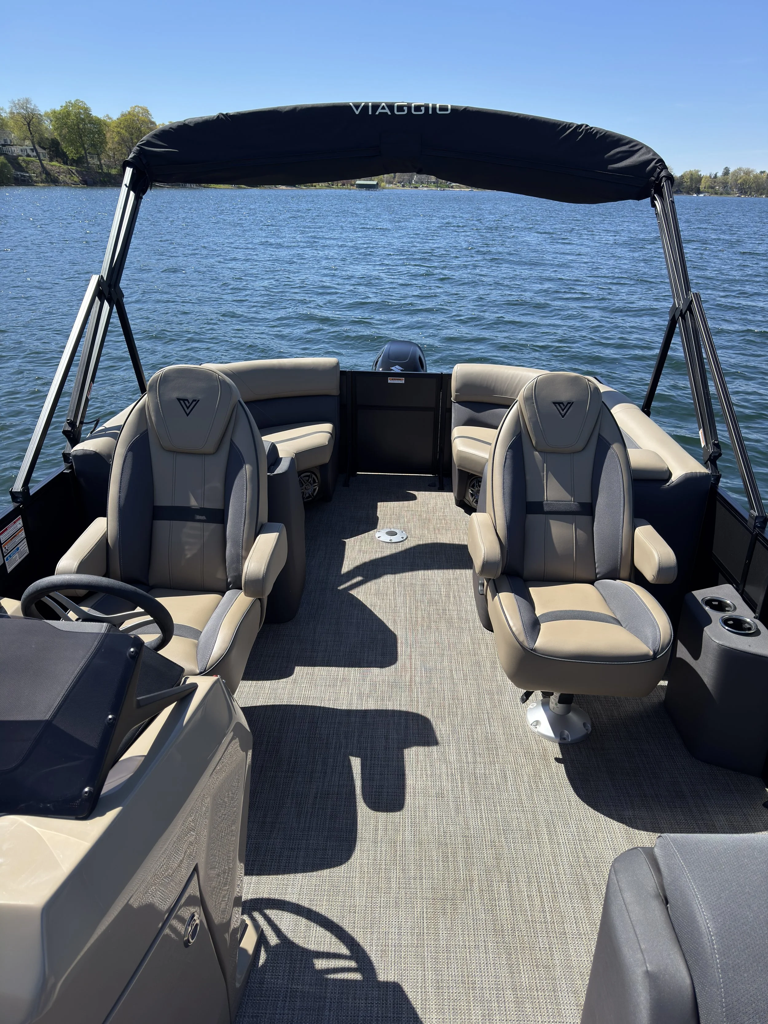 View from the front of a boat with beige and gray seating, a steering wheel, and a control panel, on a sunny day with calm water and trees in the background.