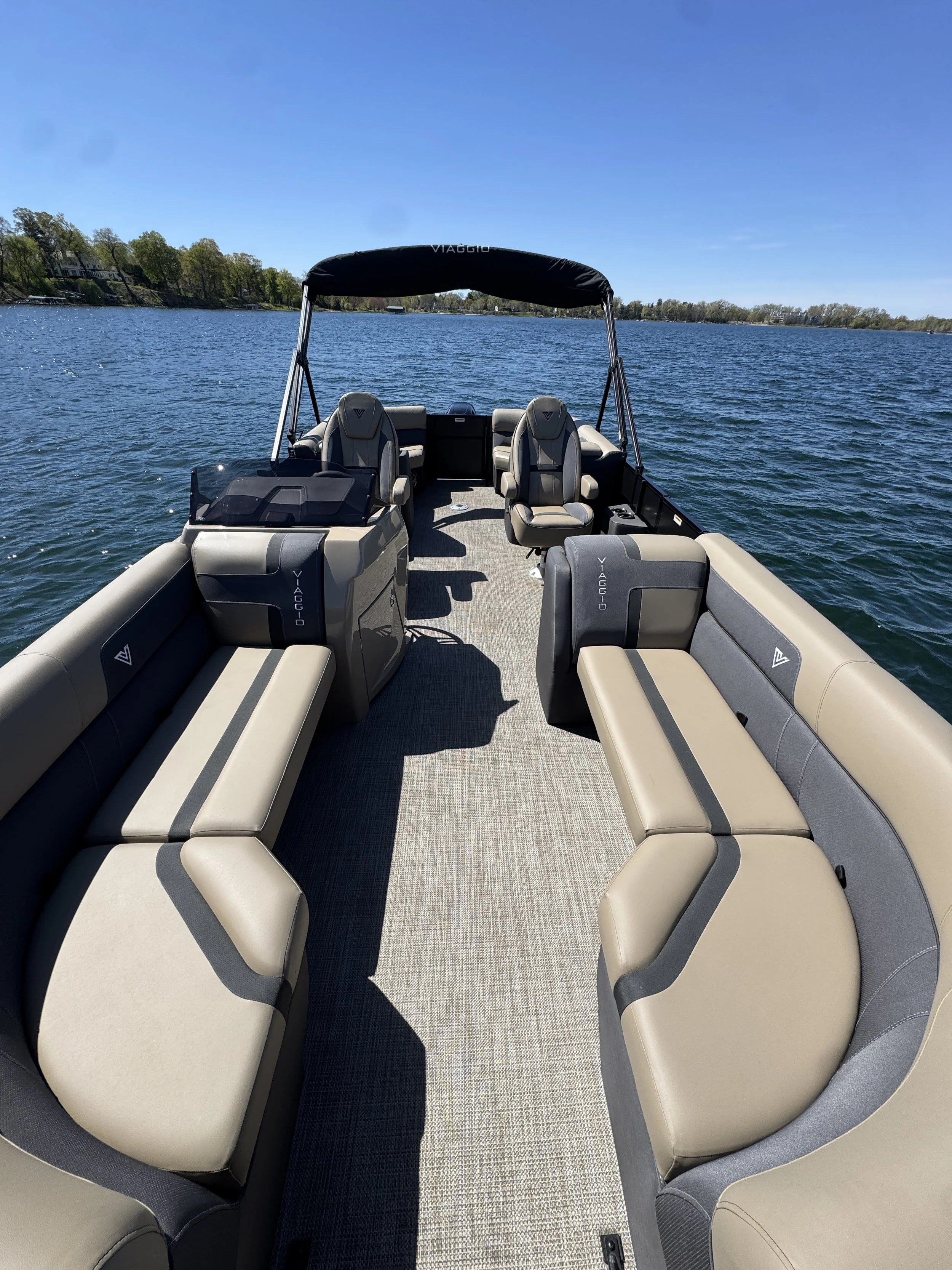 Interior of a pontoon boat on a lake with cushioned seating around the edges, a small table, and a canopy overhead under a clear blue sky.