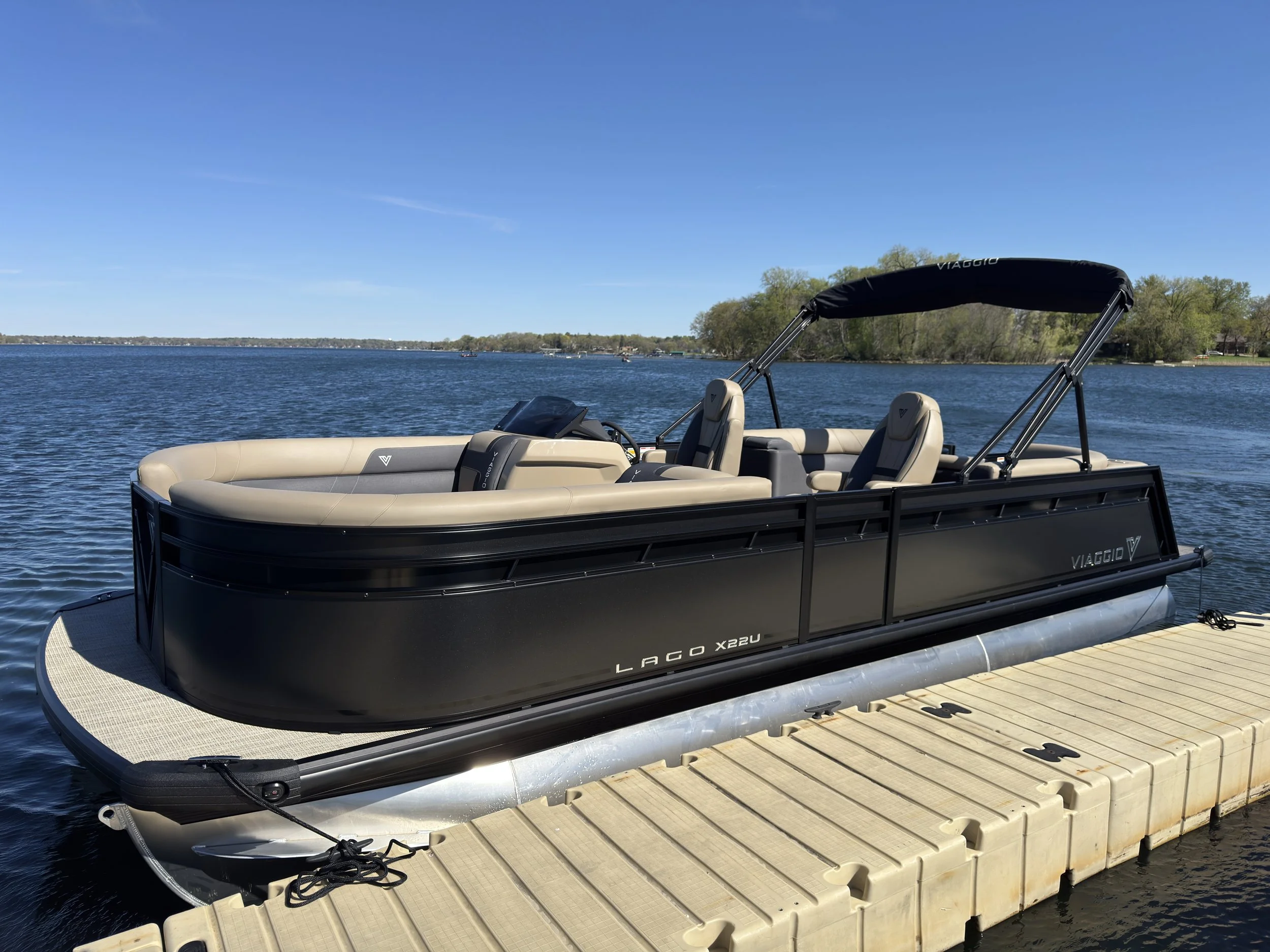 A black and beige pontoon boat docked at a wooden pier on a lake under a clear blue sky with some trees on the far shore.