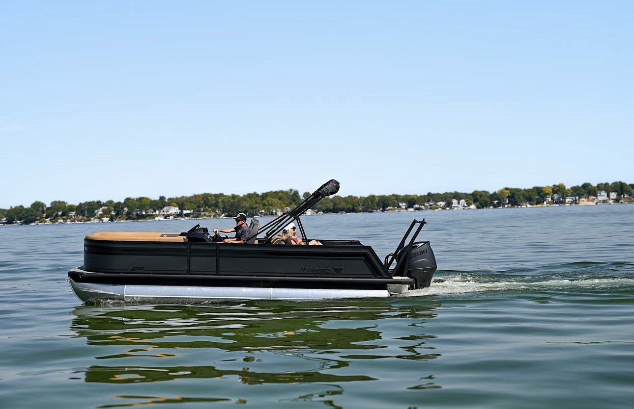 A black yacht with a beige top cruising on a lake with two people on board, one steering and the other lying down, with trees and houses in the background under a clear blue sky.