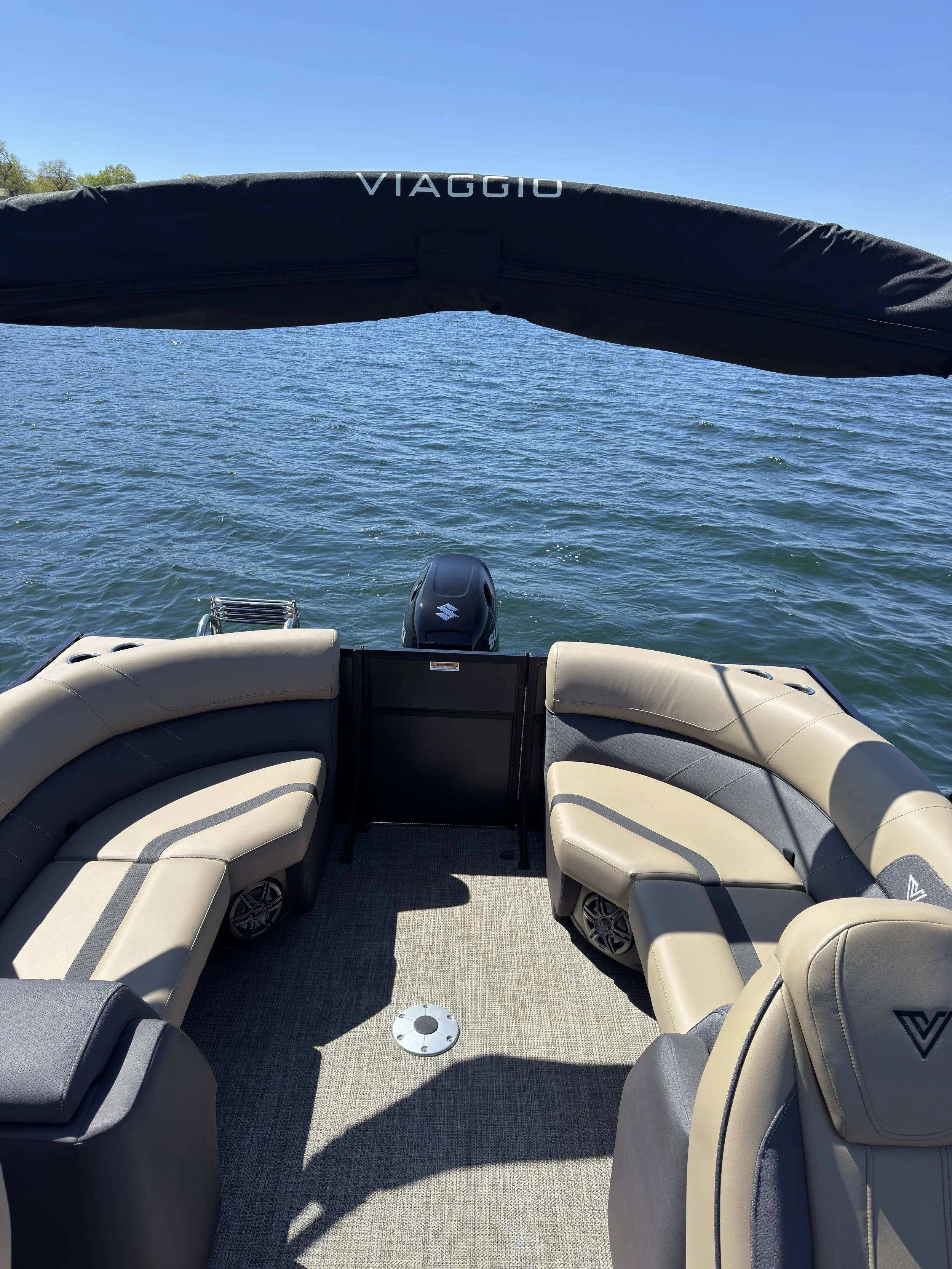 View from the back of a boat with beige and gray cushioned seats, a Suzuki outboard motor, and a black canopy with the brand name 'Viaggio'; water and a clear blue sky are visible in the background.
