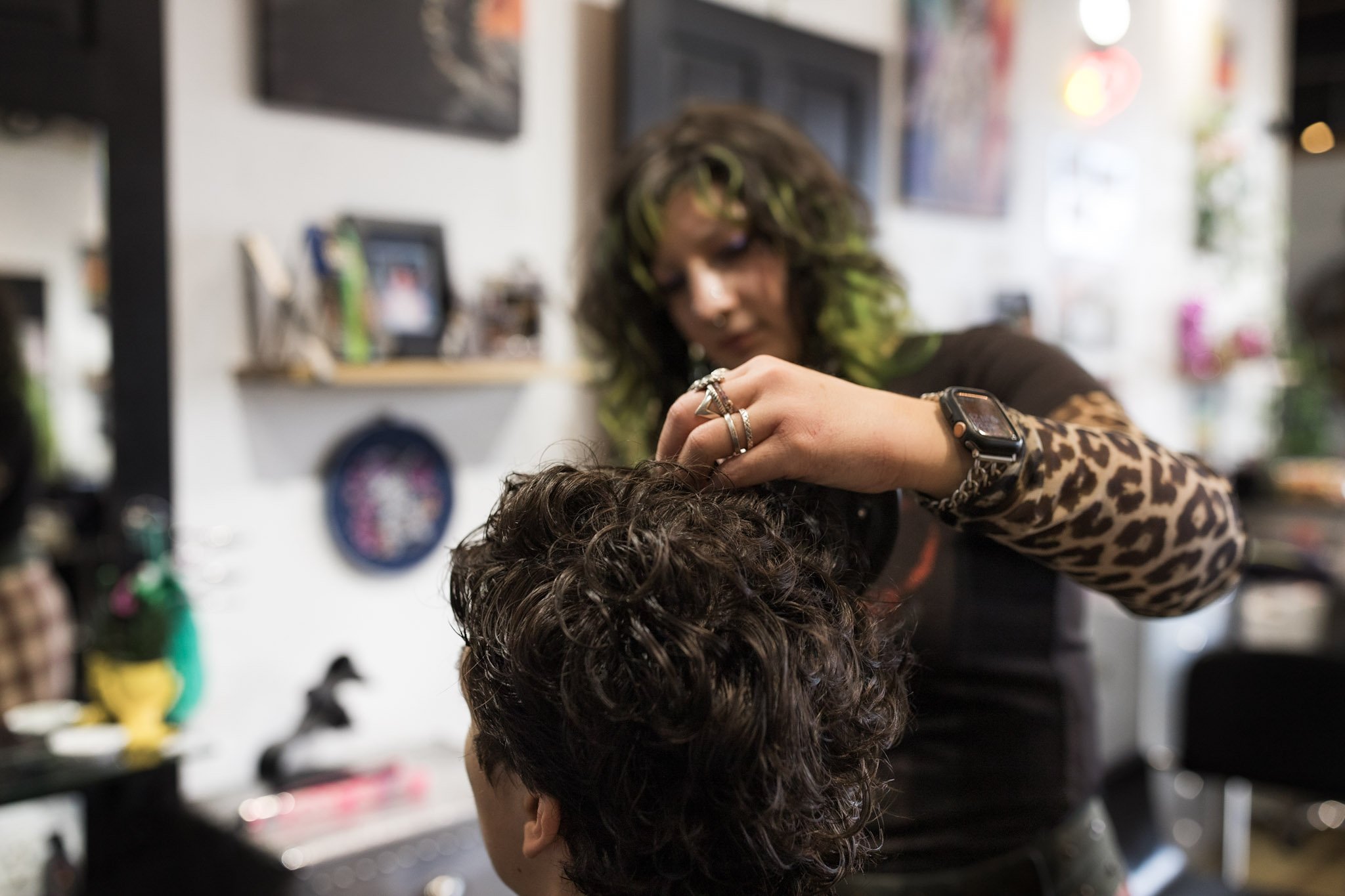 Hairdresser cutting a person's curly brown hair in a salon, with various tools and framed pictures on the wall in the background.