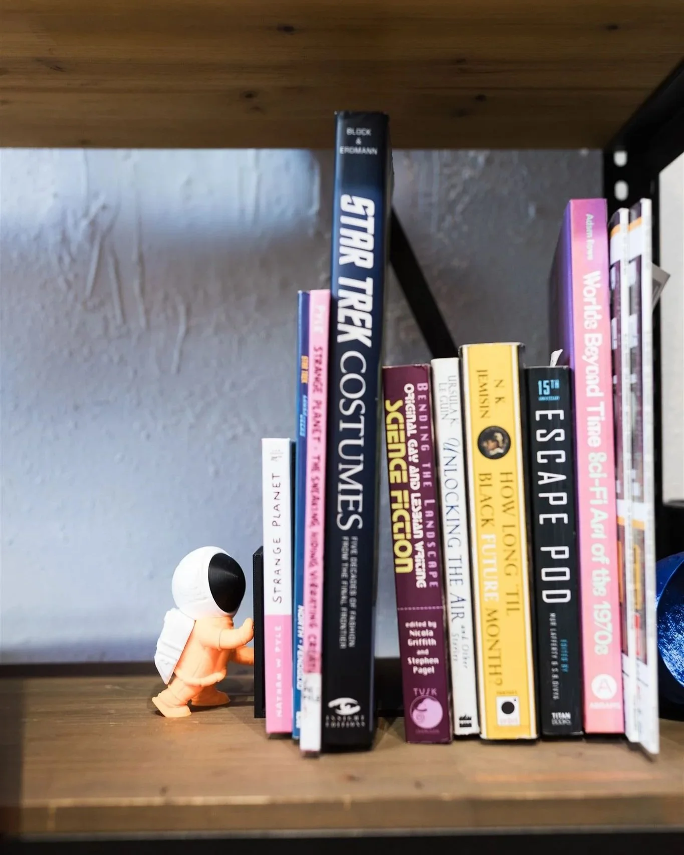 A small astronaut figurine with a white helmet and orange suit is behind a row of books on a wooden shelf.