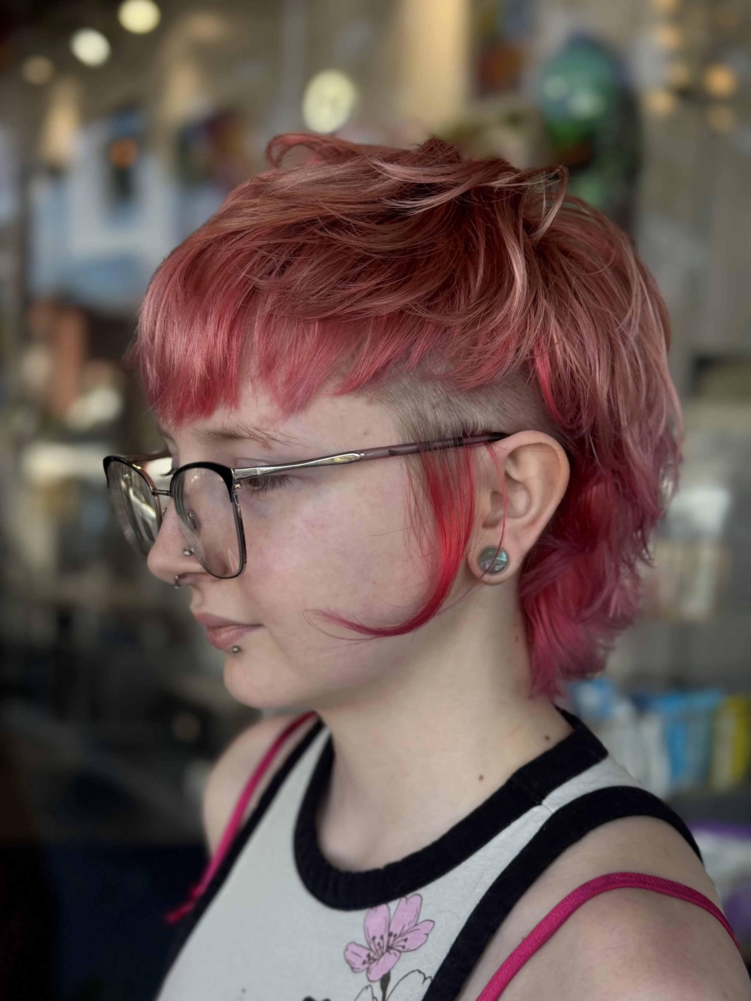 Side profile of a person with short, layered, pink hair and glasses, wearing a sleeveless top with floral design and black and white stripes on the collar, in an indoor setting with blurred colorful background.